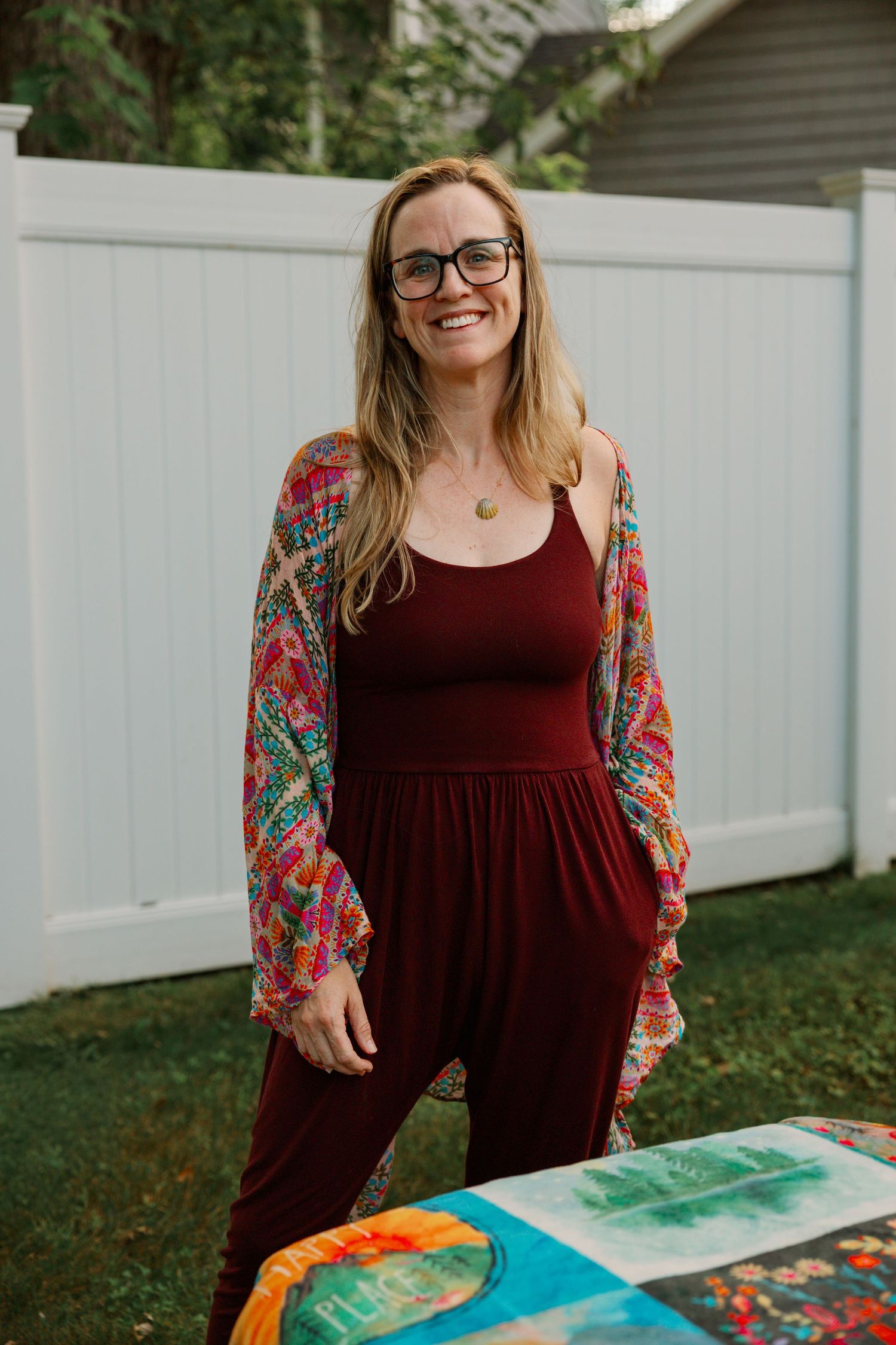 Smiling woman with glasses wearing a burgundy jumpsuit and colorful floral kimono standing in a backyard.