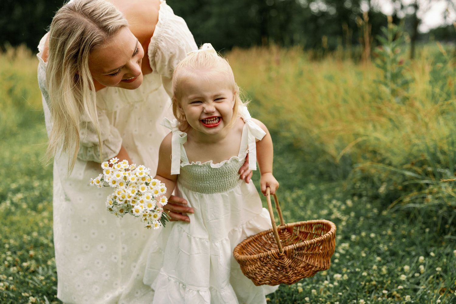Laughing toddler girl in white dress holds a basket while mom holds wildflowers outdoors.