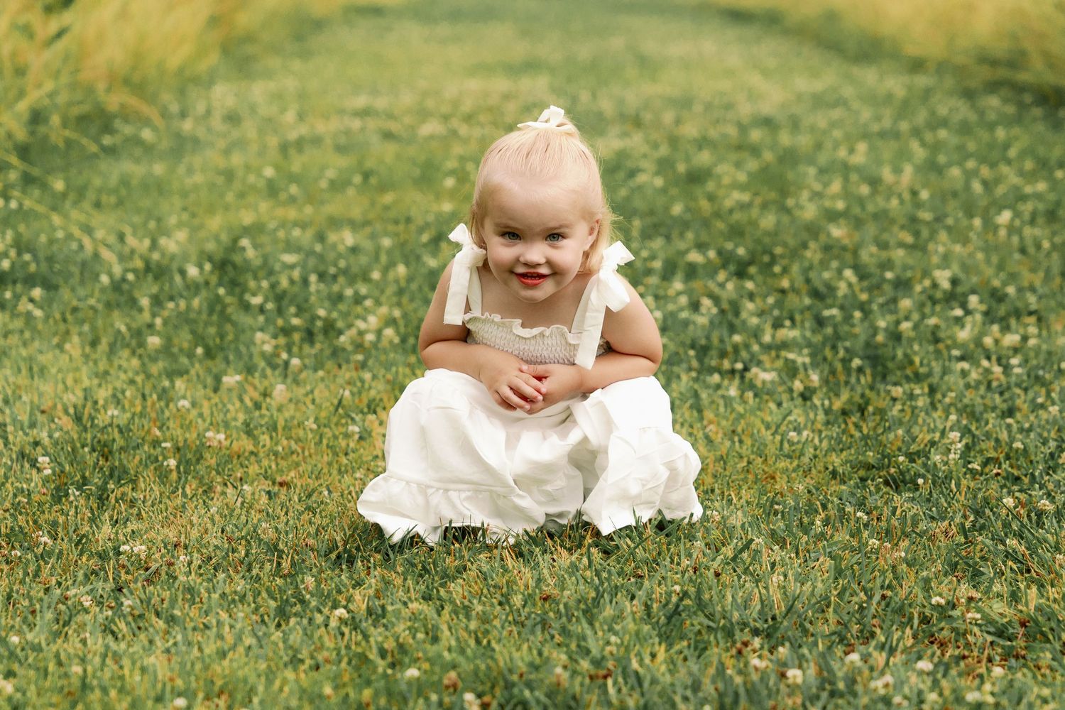 Cute blonde toddler girl in white dress sits cross-legged smiling on a green grassy field.
