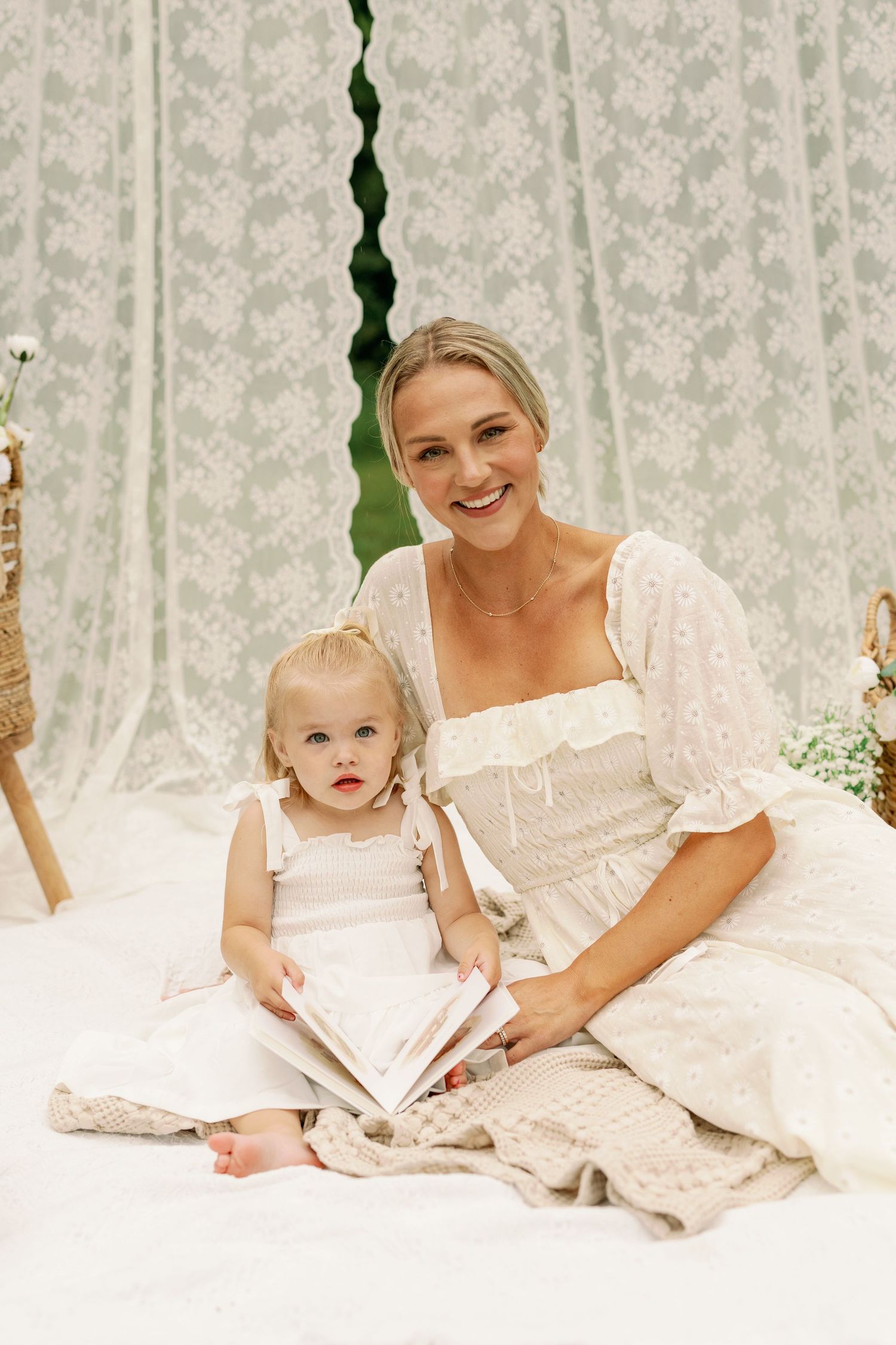 Mother and toddler daughter in matching white dresses reading a book together on a blanket.