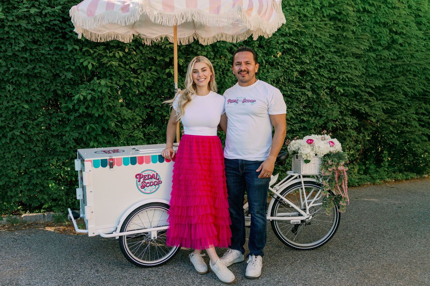 Smiling couple standing together beside a white ice cream bicycle cart with a fringed umbrella.