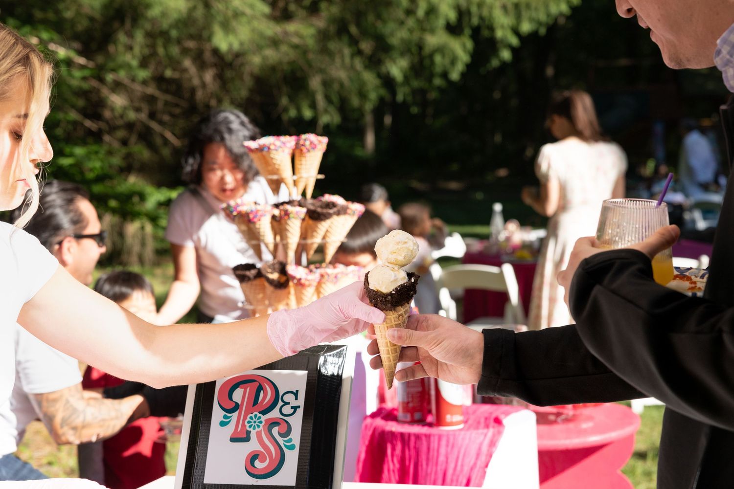 People serving ice cream cones from a colorful display tower at an outdoor event with pink decorations.