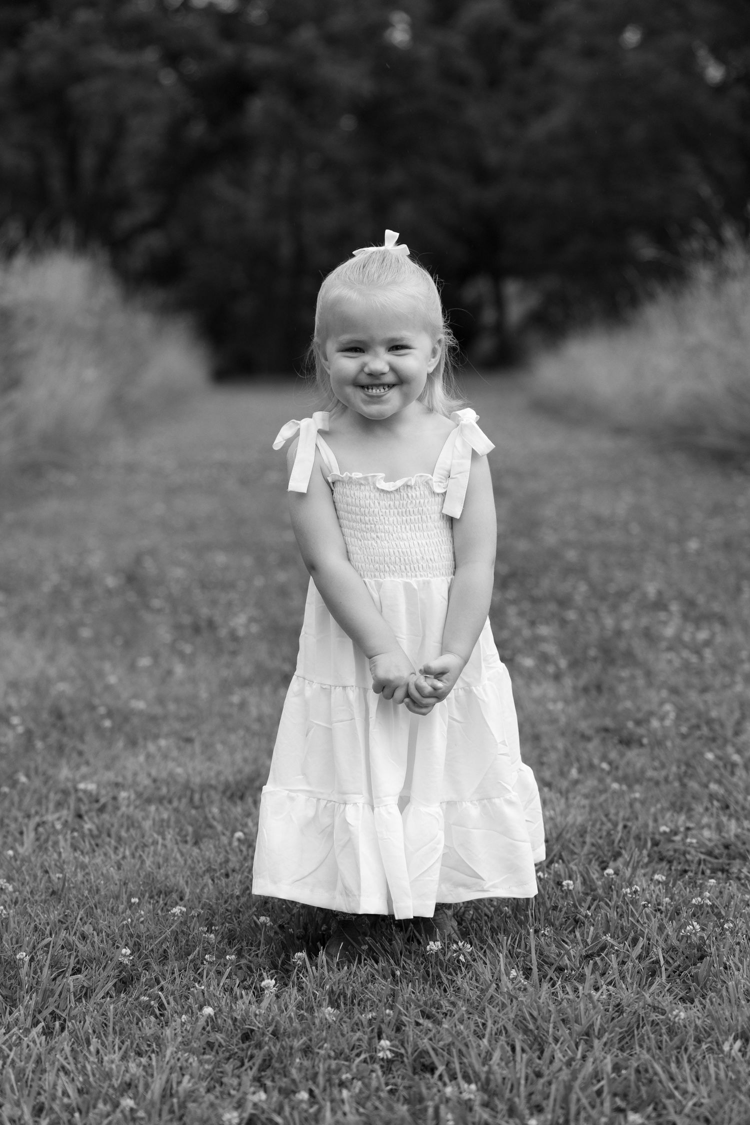 Black and white portrait of a smiling blonde toddler girl in white dress standing in a park.