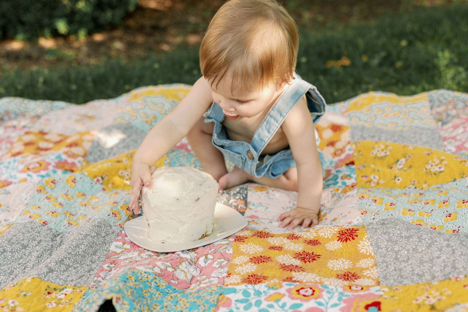Baby girl in denim romper playing with sun hat on colorful patchwork quilt outdoors.