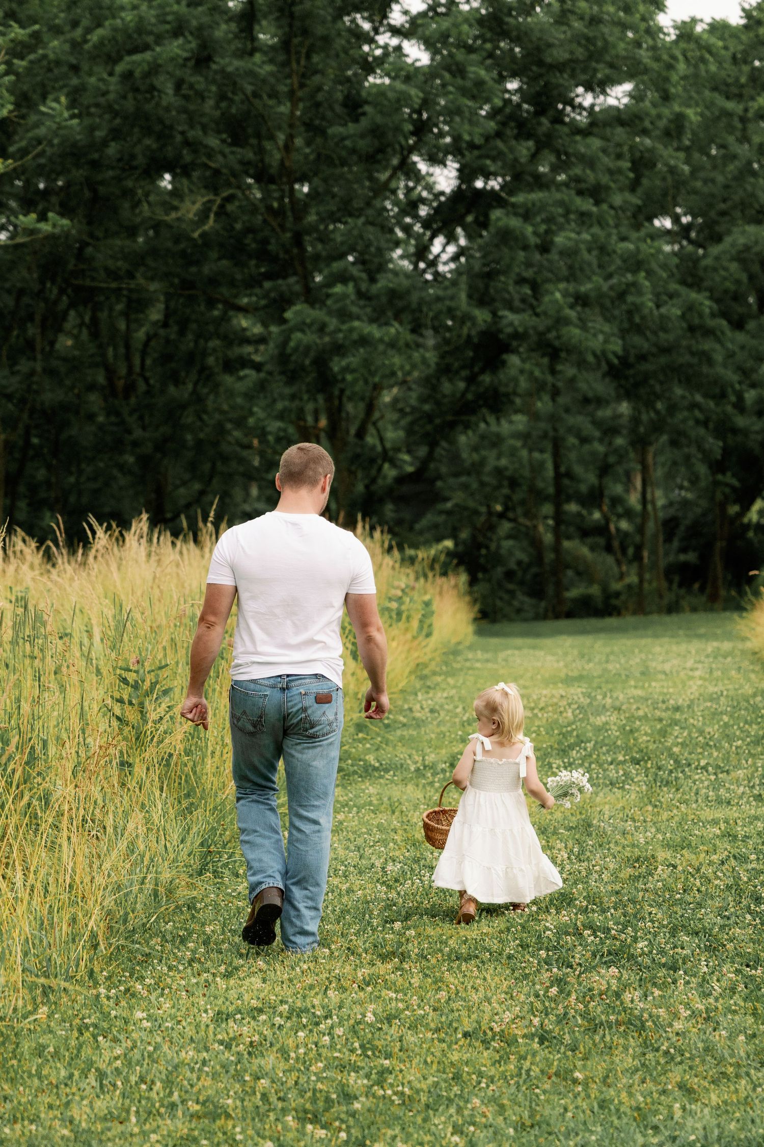 Father in white t-shirt and jeans walking with young daughter carrying a basket in a lush green field.