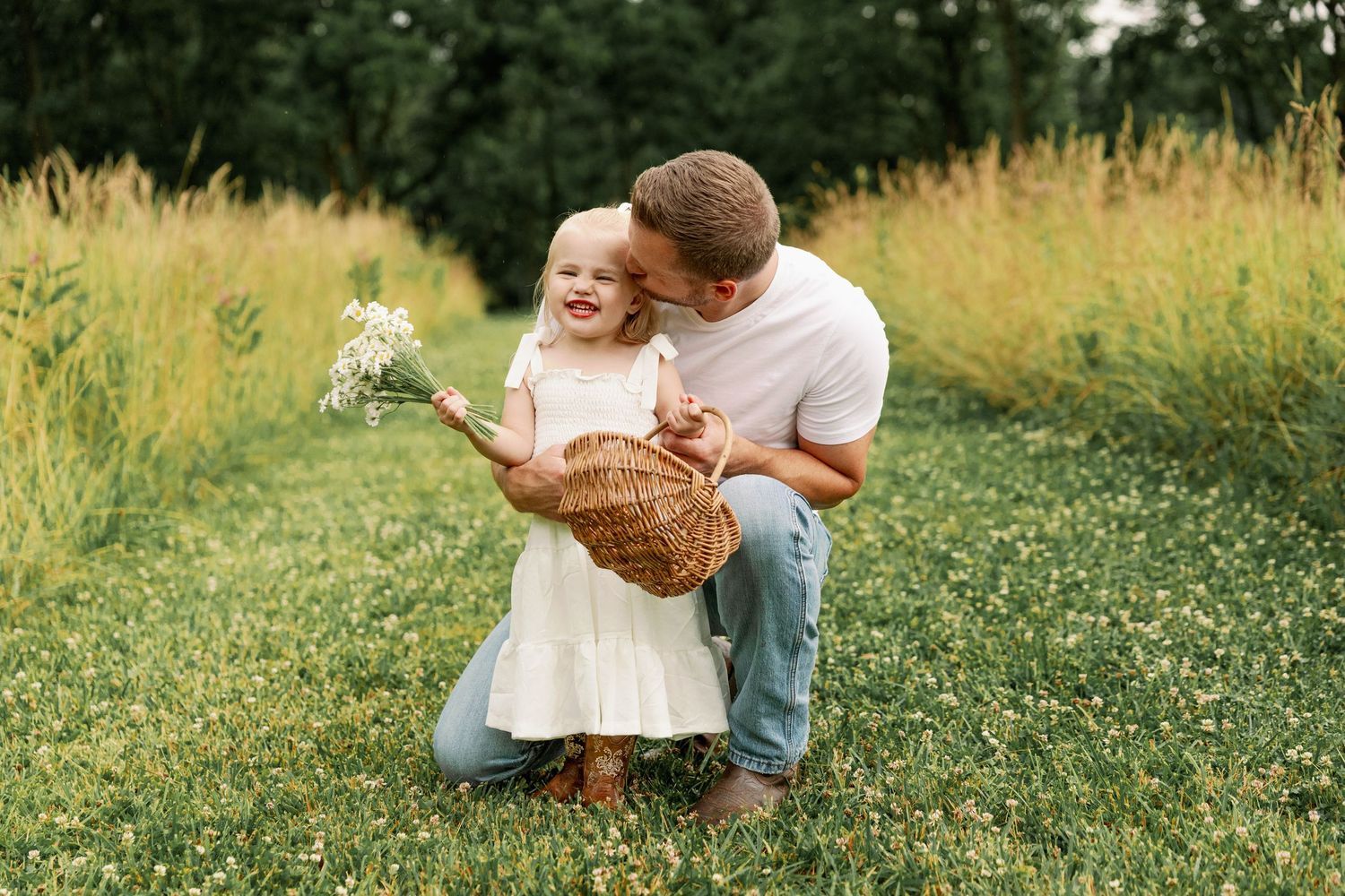 Loving father kneels to kiss smiling toddler daughter holding wildflowers and wicker basket in a green grassy field.