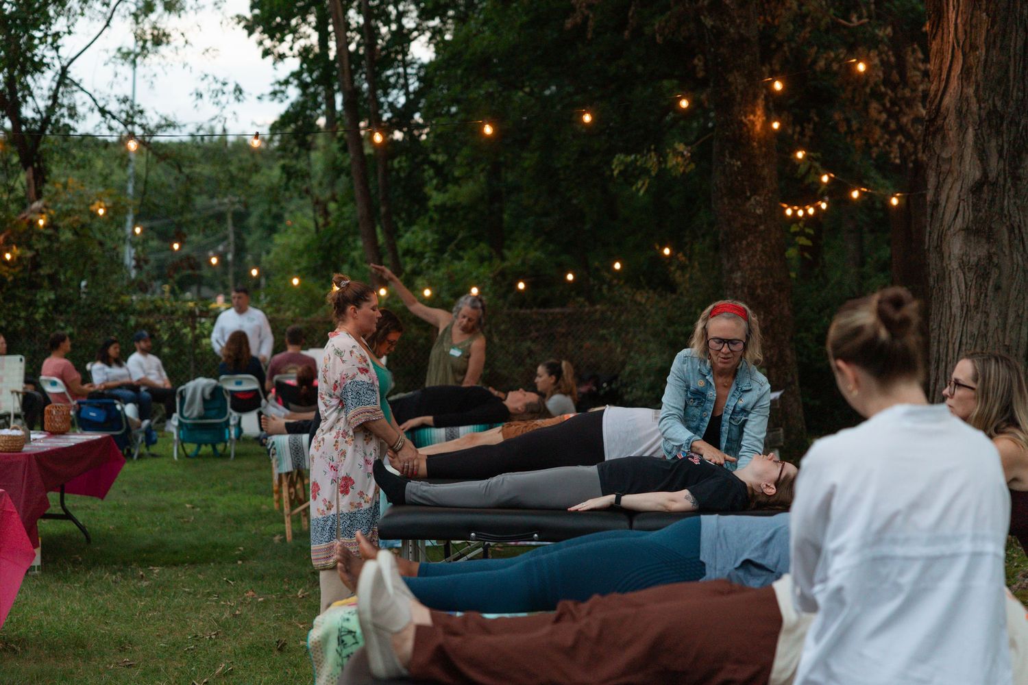 Outdoor evening massage event under string lights in a lush garden with therapists treating guests on tables.