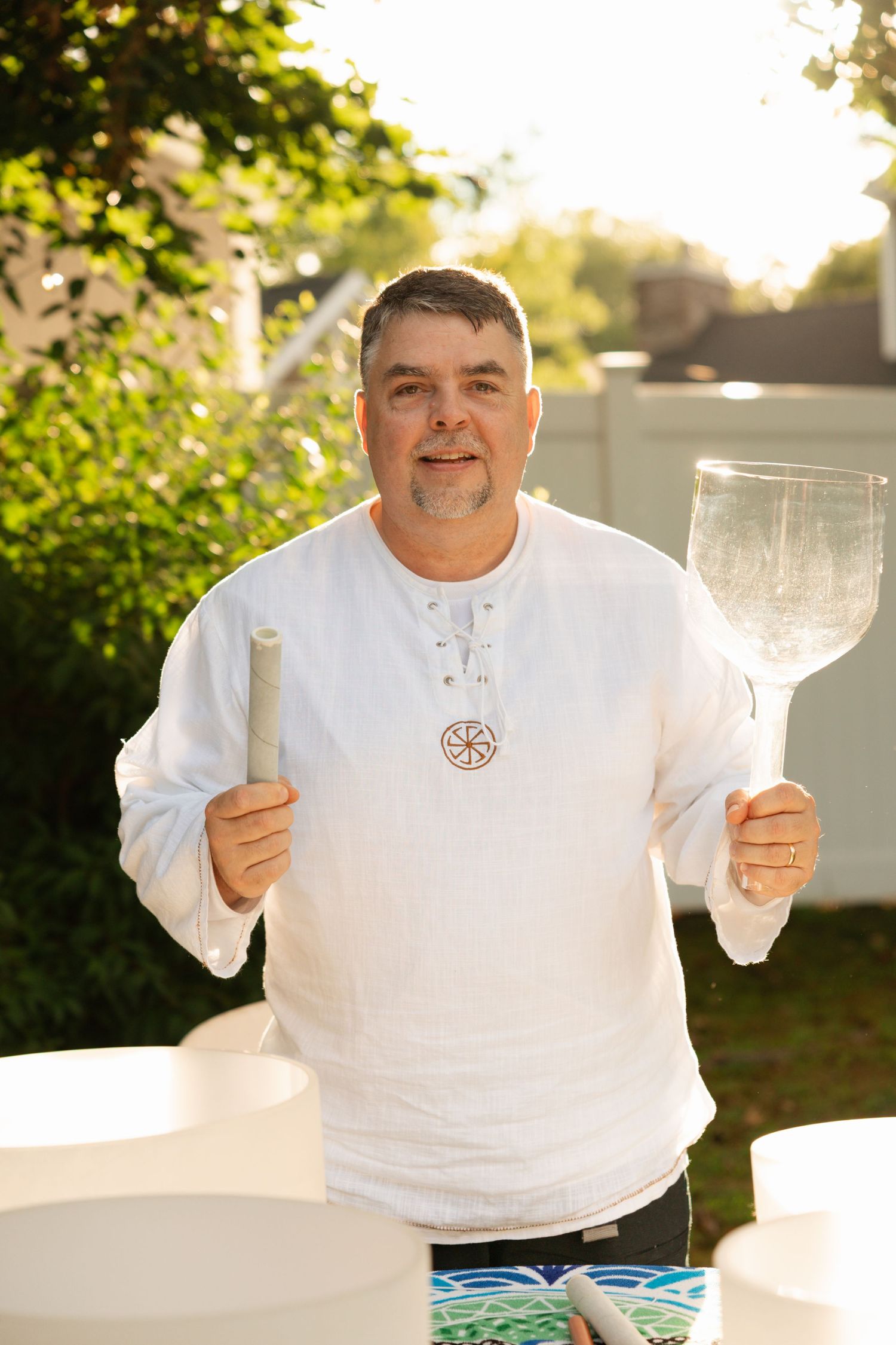 Smiling man in white linen shirt holding crystal singing bowls and mallet outdoors in sunlight.