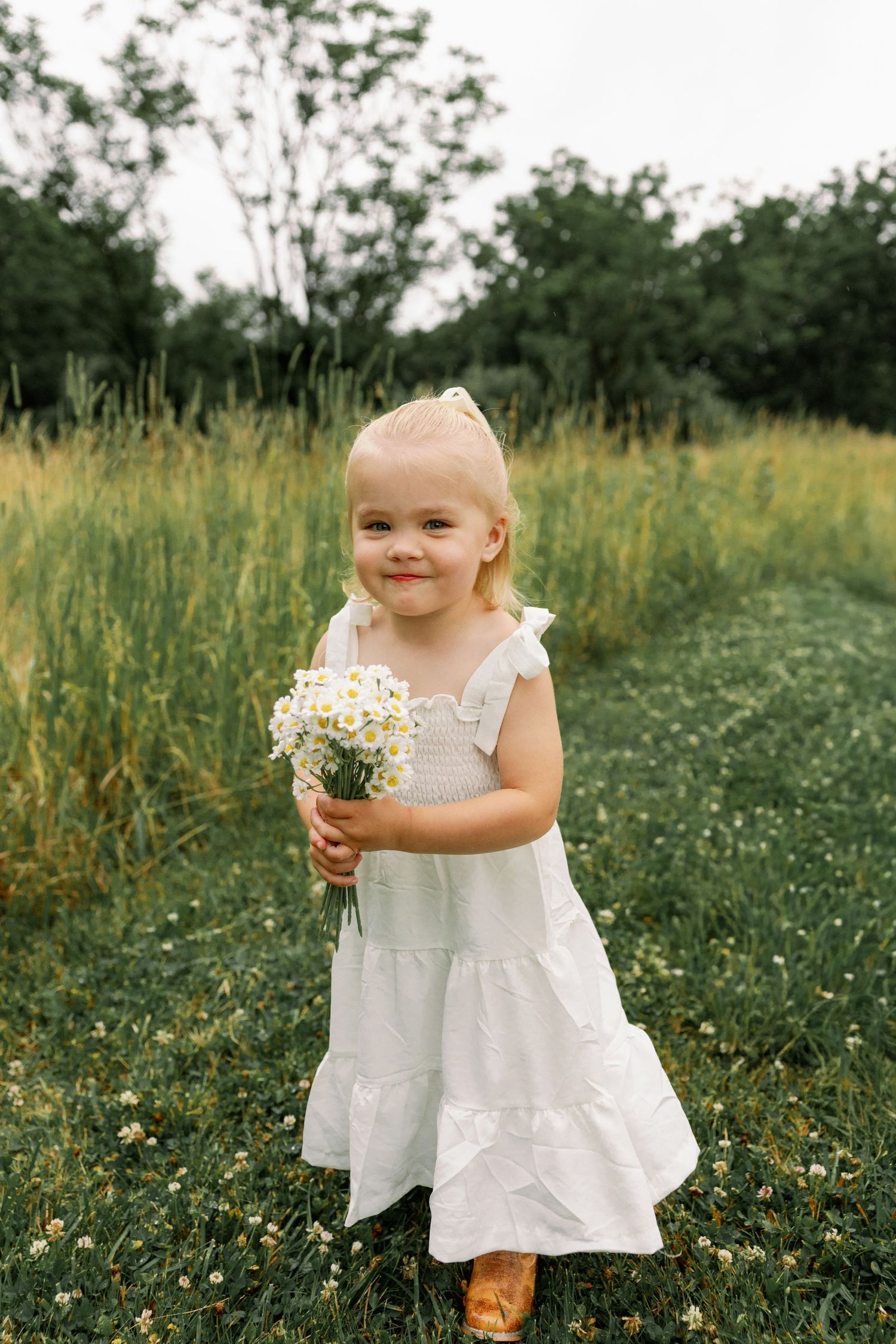 Sweet little blonde girl in white dress holds wildflower bouquet while smiling in a green meadow.