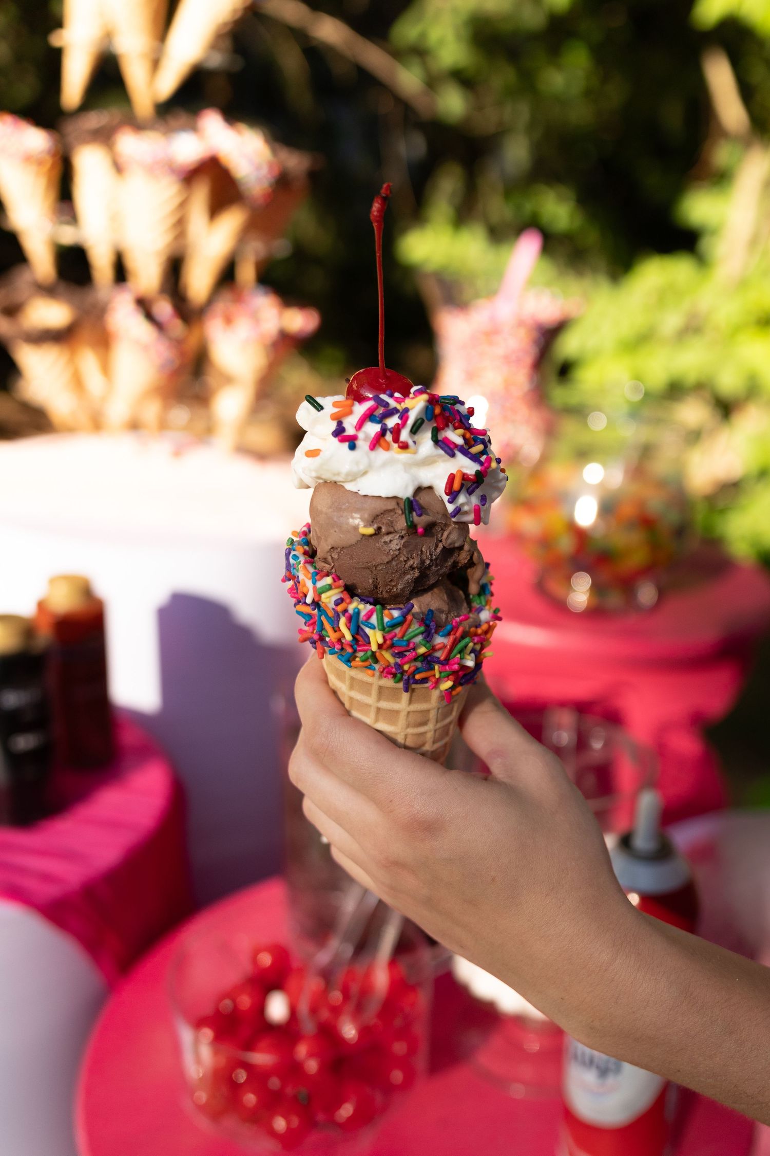 Hand holding a chocolate ice cream waffle cone with rainbow sprinkles, whipped cream, and a cherry on top.