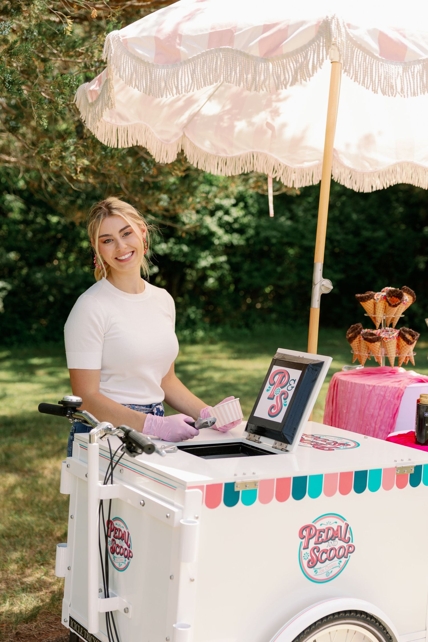 Smiling woman serving ice cream at a Pedal Scoop cart with a fringed umbrella outdoors.