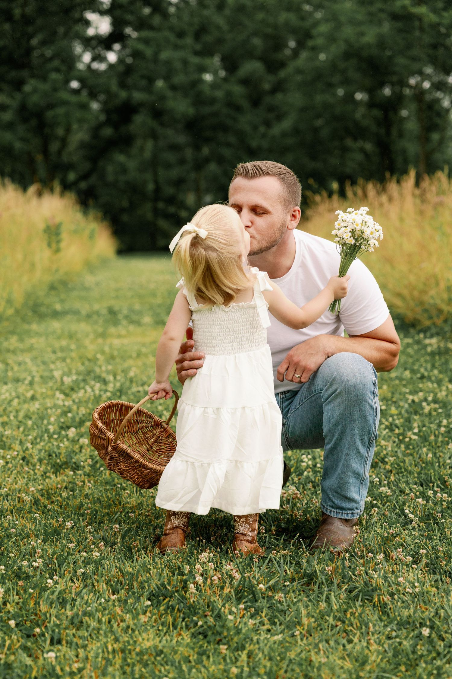 Father in white shirt kisses toddler daughter holding wildflowers and wicker basket in green field.