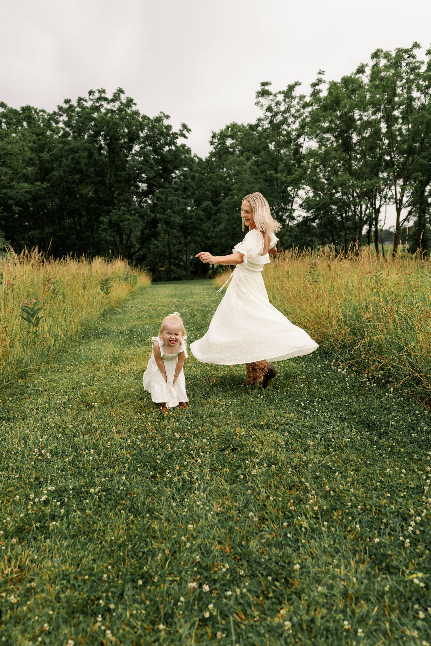 Mother and toddler daughter in matching white dresses twirl playfully in a green field.