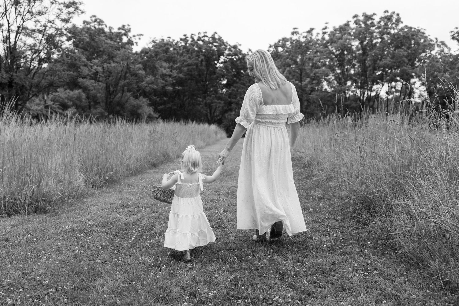 Black and white photo of mother and daughter in white dresses walking hand in hand along a grassy field path.