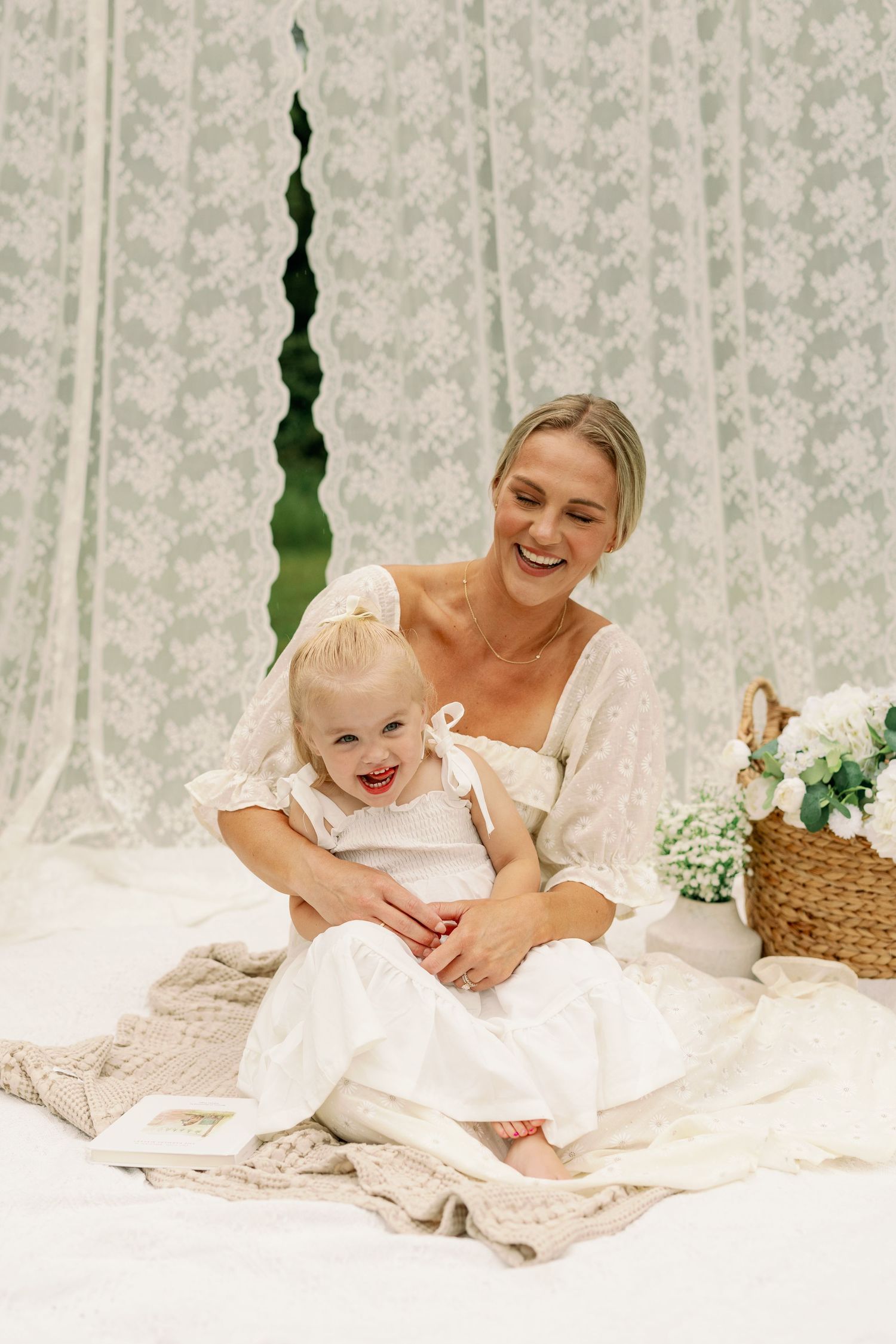 Laughing mother in white dress holding giggling toddler on blanket with lace backdrop and flowers.