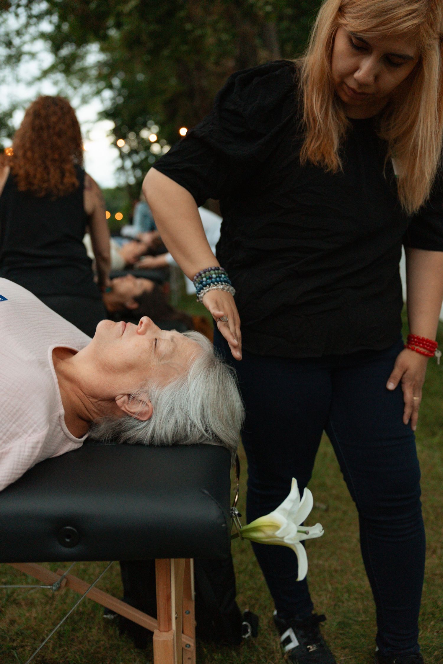 A massage therapist performs a relaxing head massage on an elderly woman lying on a portable table outdoors.