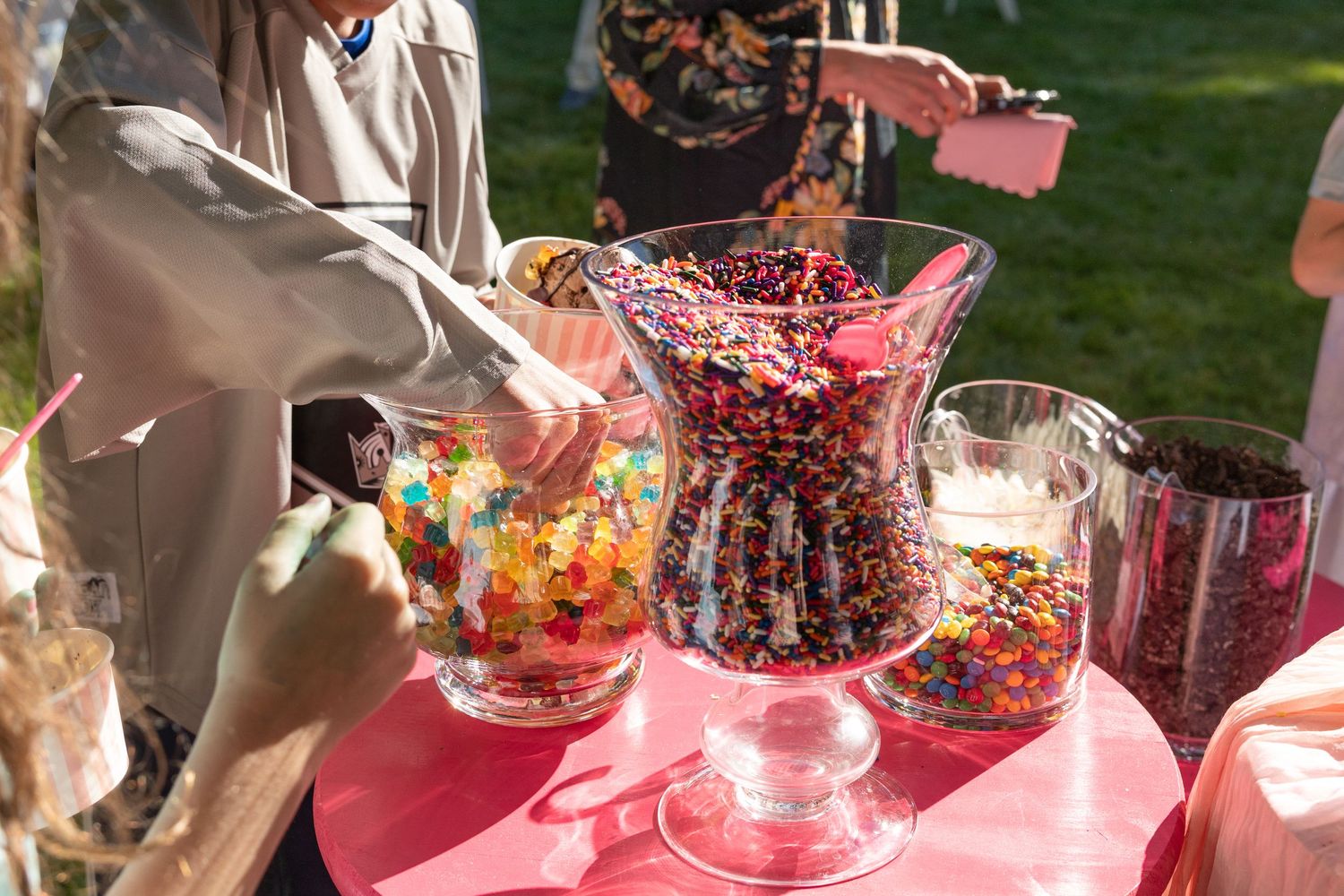 Person scooping colorful sprinkles from large glass bowls at an outdoor ice cream topping bar.