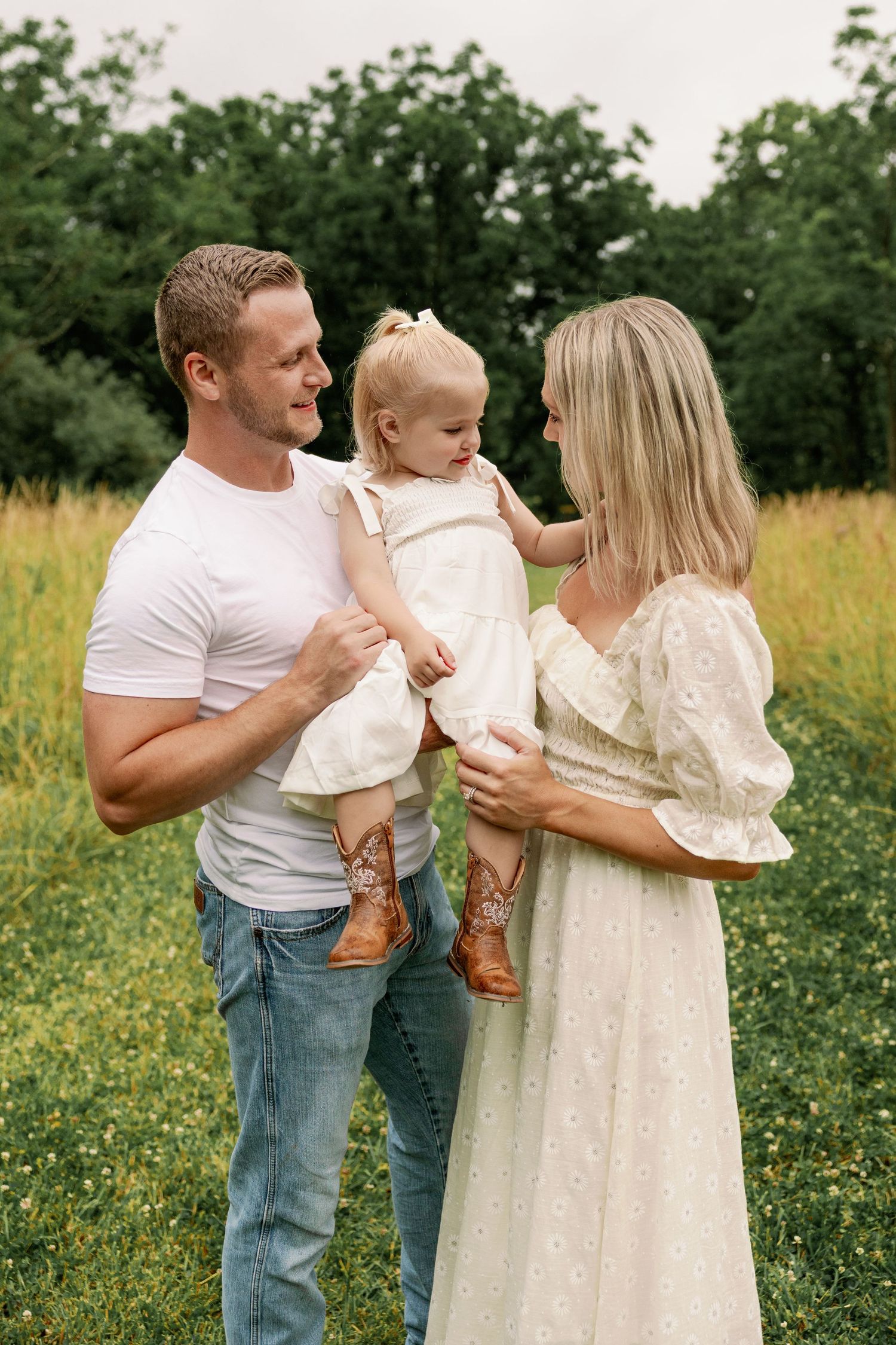 Happy family of three standing in a green meadow, parents smiling at their toddler daughter.