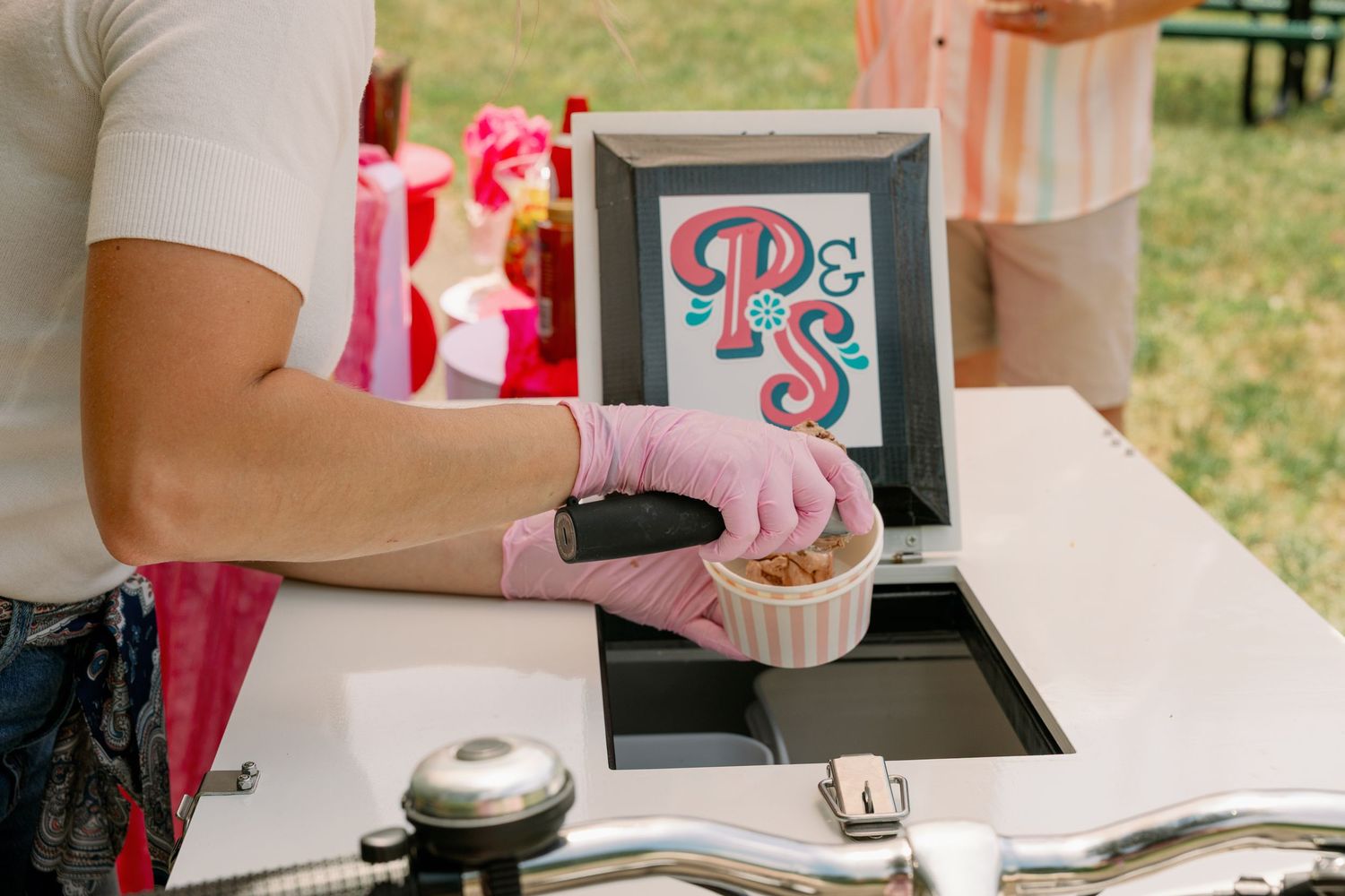 Gloved hands scooping ice cream at a Pedal Scoop cart with a monogram sign displayed.