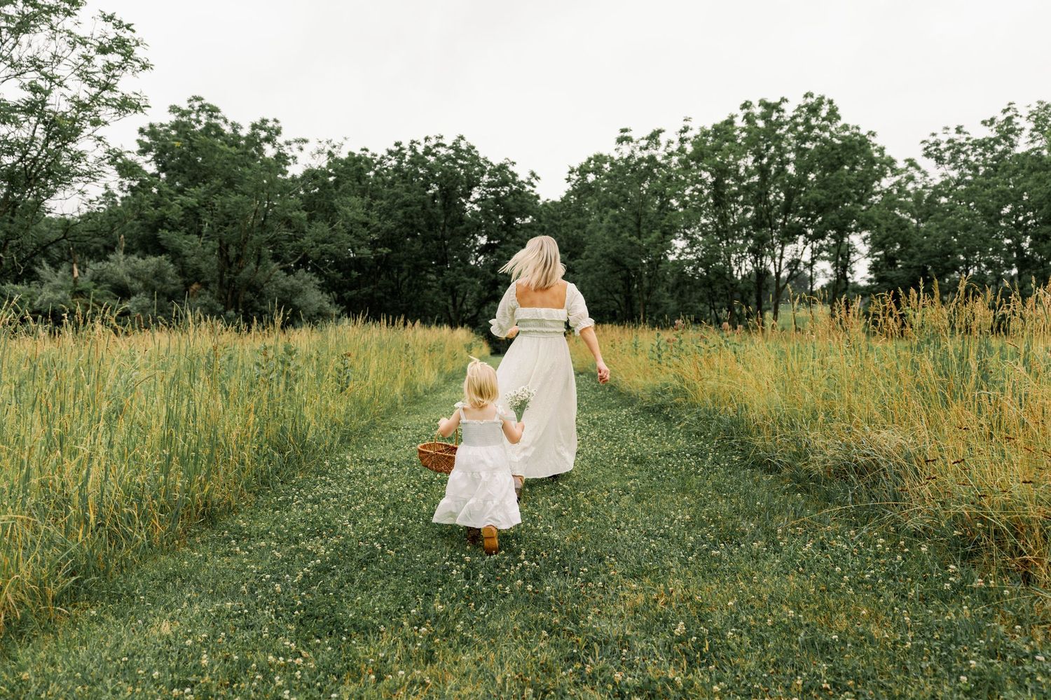 Mother and toddler daughter in white dresses walk hand in hand through a green meadow.