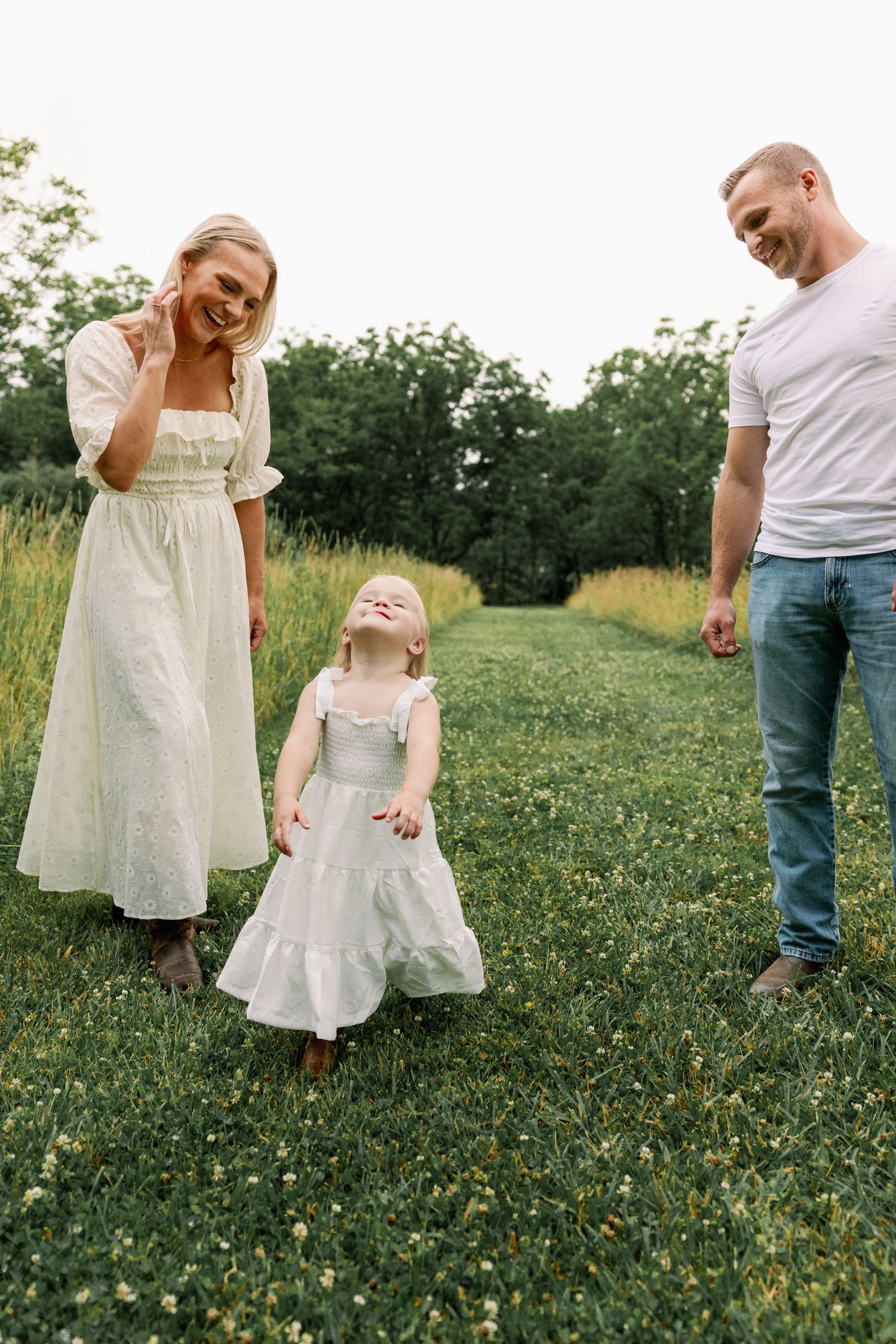 Happy family with smiling parents watching their toddler daughter twirl in a white dress on a green lawn.