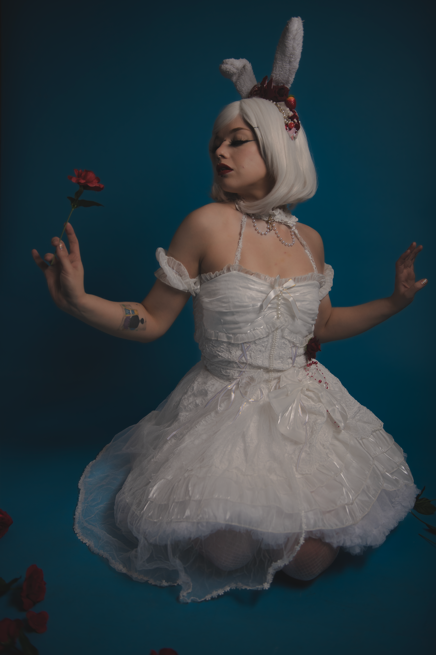 A dreamy fashion photo of a model in a white tulle dress and bunny ears posing with red roses against a blue backdrop.