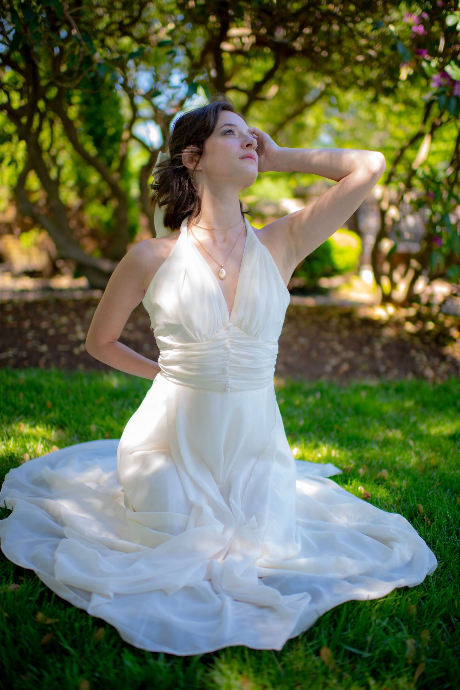 Elegant white wedding dress flowing on green grass in a sunlit garden setting with lush foliage backdrop.