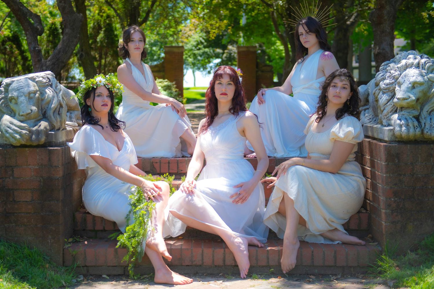 A group in flowing white dresses pose together on stone steps in a garden setting with greenery and sunlight.