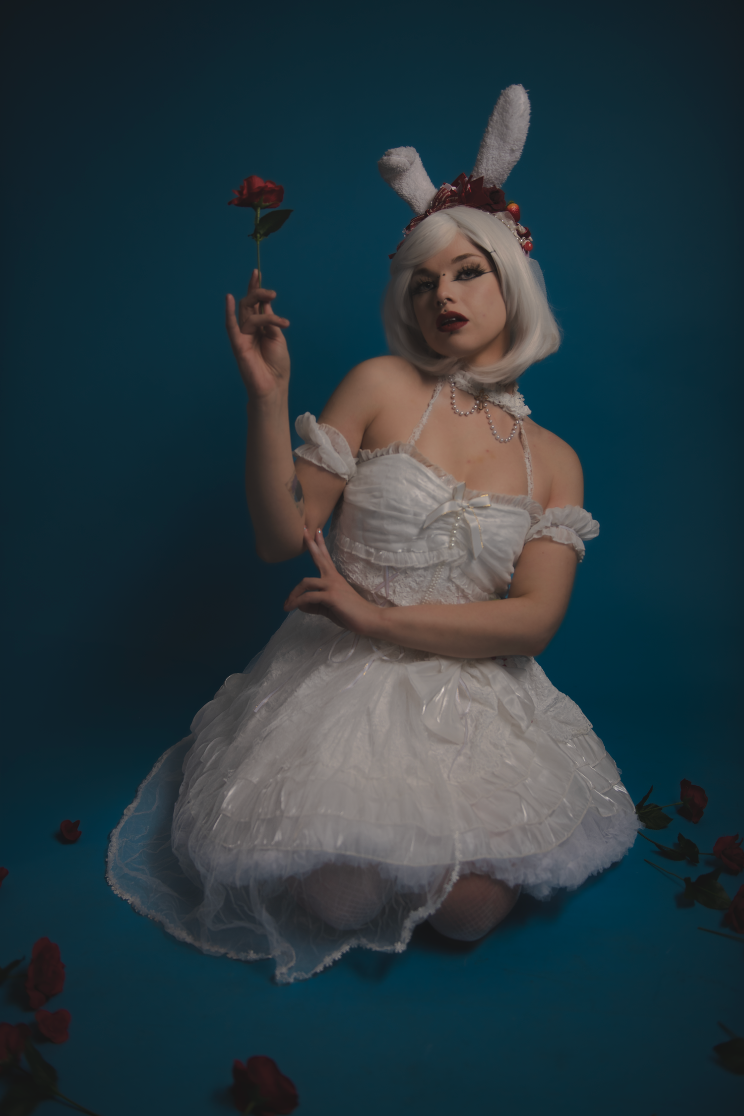 A figure in a white tulle dress and bunny ears holds a red rose against a dark teal background.