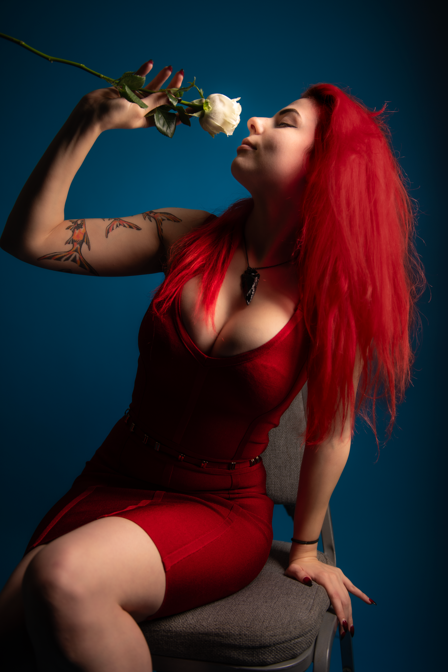 A figure in a red dress with vibrant red hair poses with a white rose against a dark blue background.