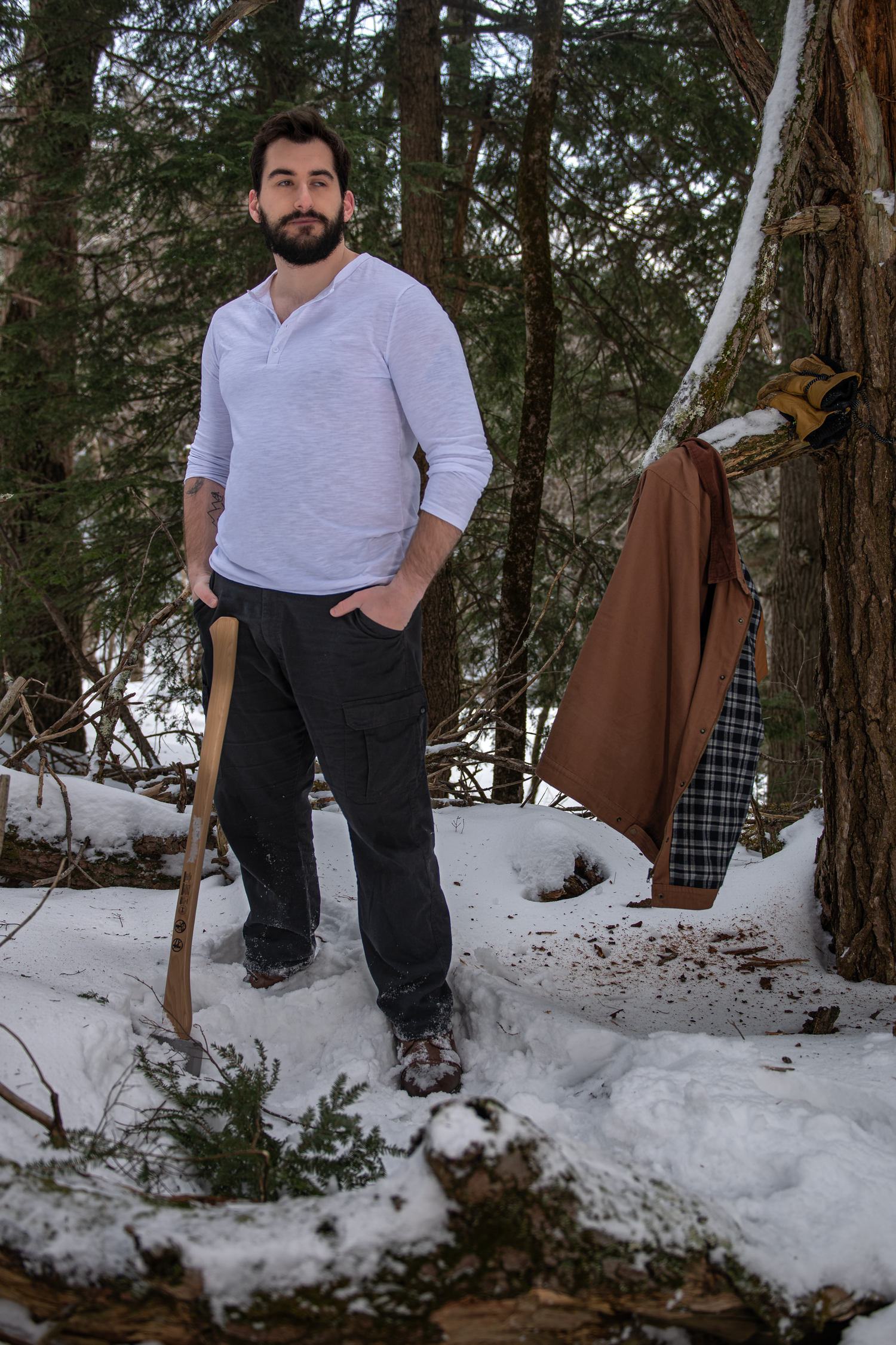 A person in a light purple henley shirt stands in snowy woods holding a walking stick while winter clothing hangs nearby.