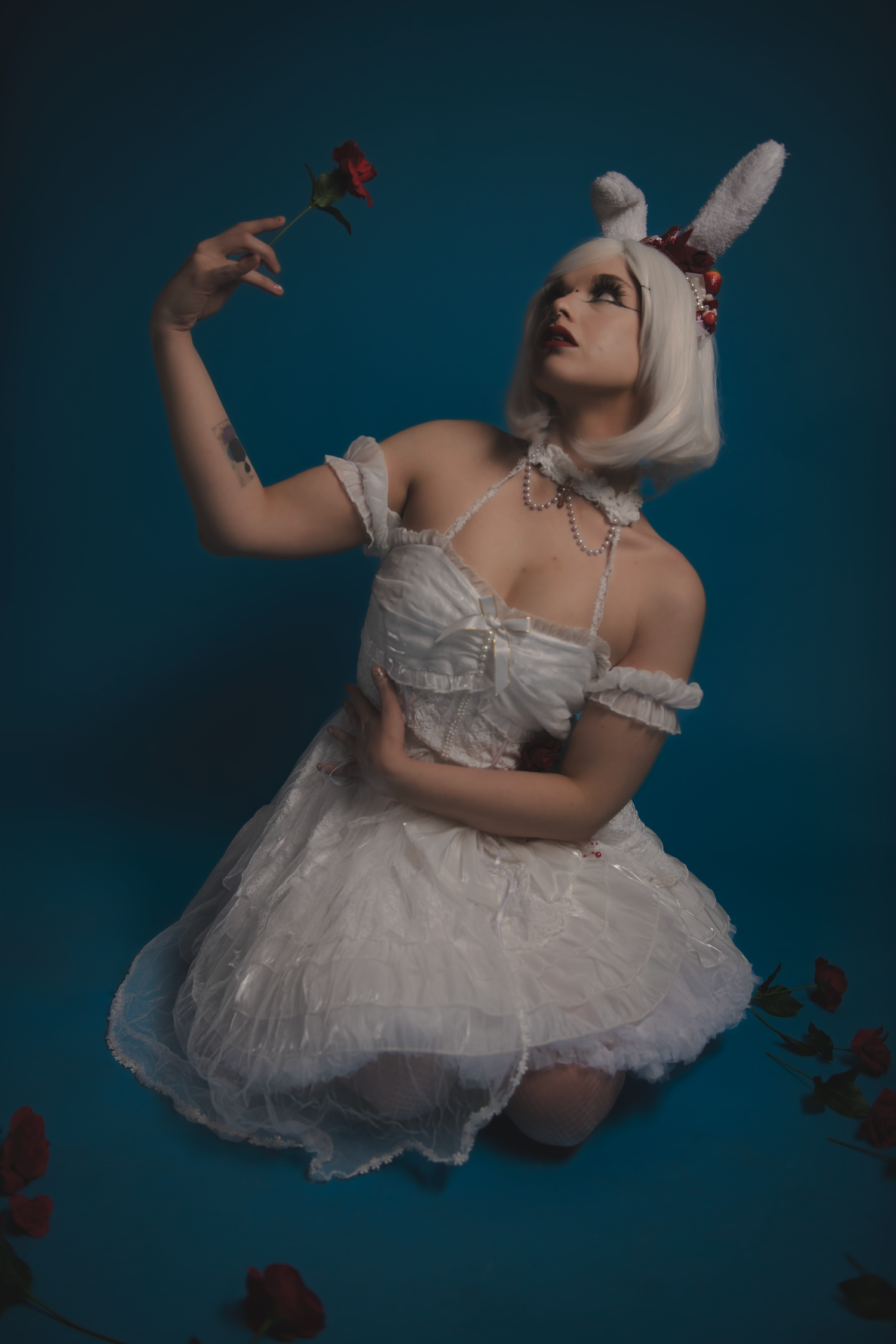 A figure in a white lace costume with bunny ears holds a red rose while posing against a dark teal background.