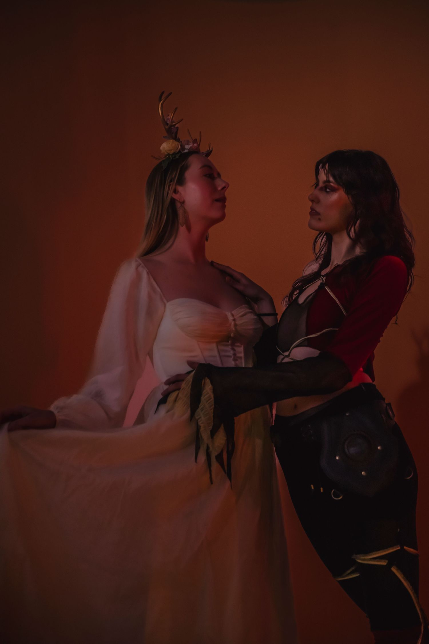 Two women in contrasting dark and light outfits pose dramatically against a moody reddish-brown backdrop.