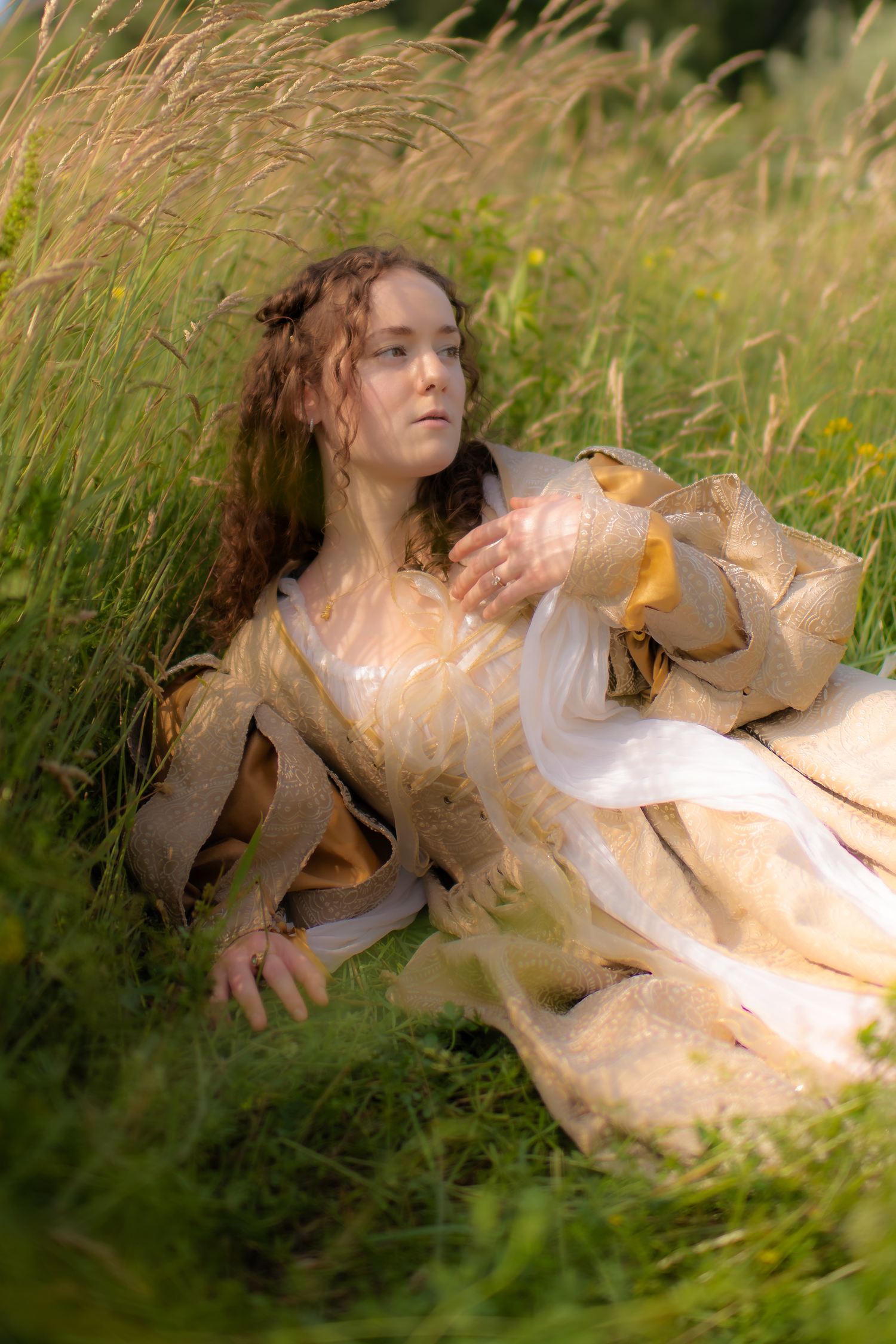 A dreamy portrait of a person in vintage-style dress lying in tall grass during golden hour.