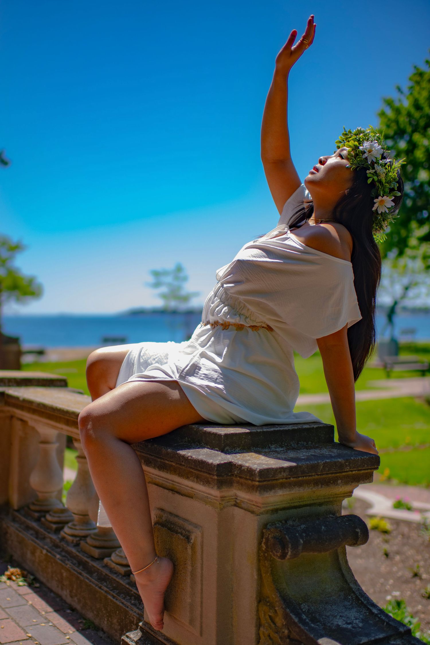 Person in white dress poses gracefully on stone ledge against blue sky and ocean backdrop on sunny day.