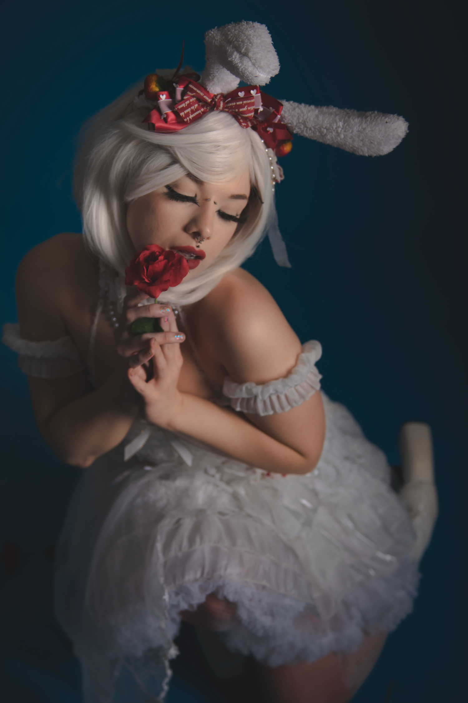 Artistic portrait featuring white dress, plush bunny ears and red rose against moody dark backdrop.