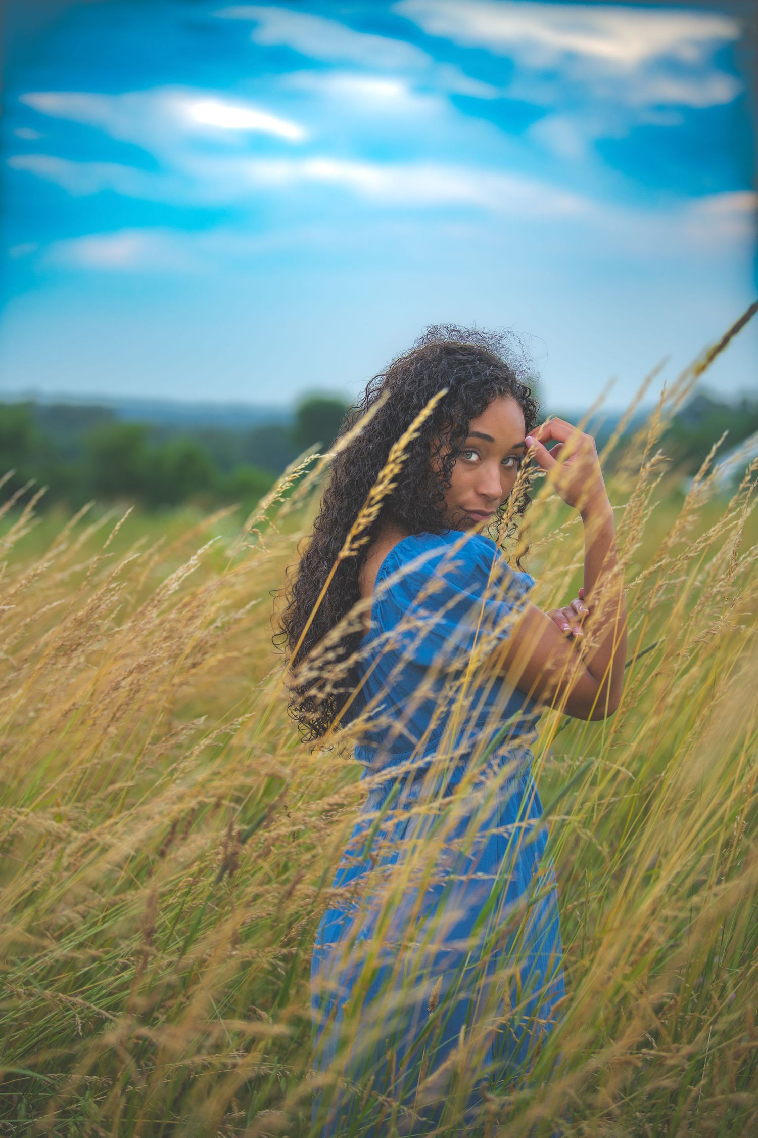 Woman in flowing blue dress poses artistically among golden wheat fields.