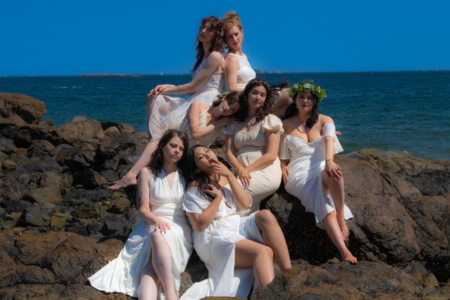 A group in white dresses poses dramatically on coastal rocks against a deep blue ocean backdrop.
