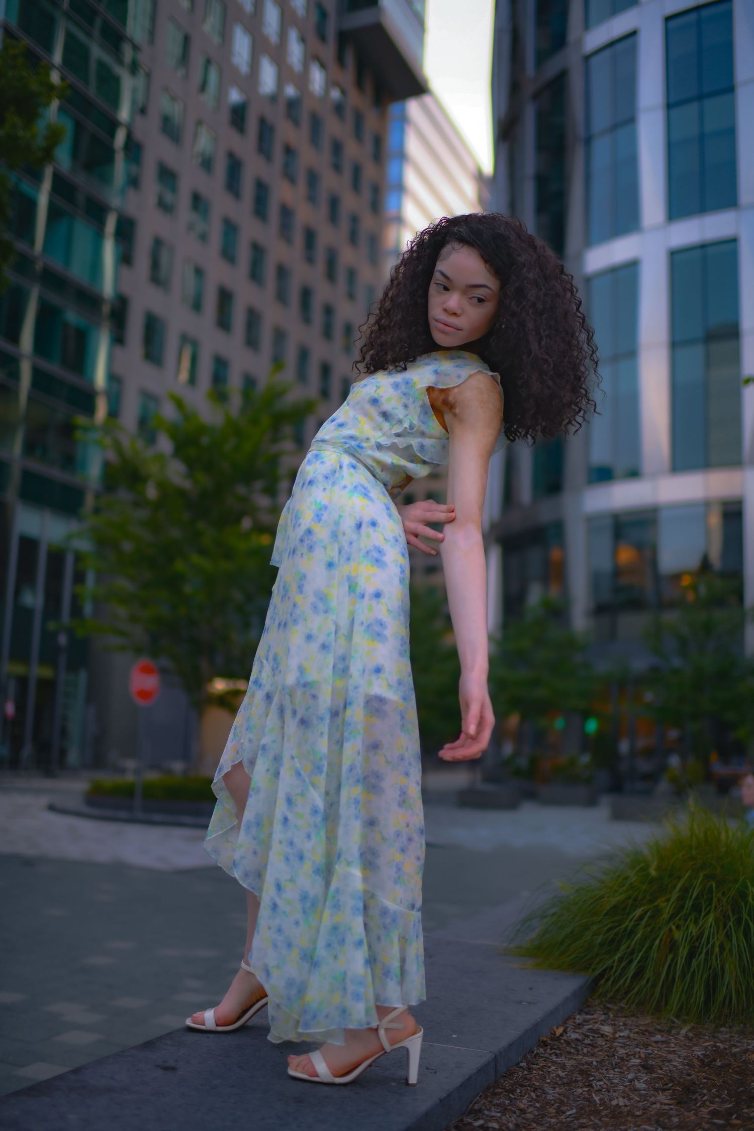 A floral dress and curly hair against modern city buildings at dusk.