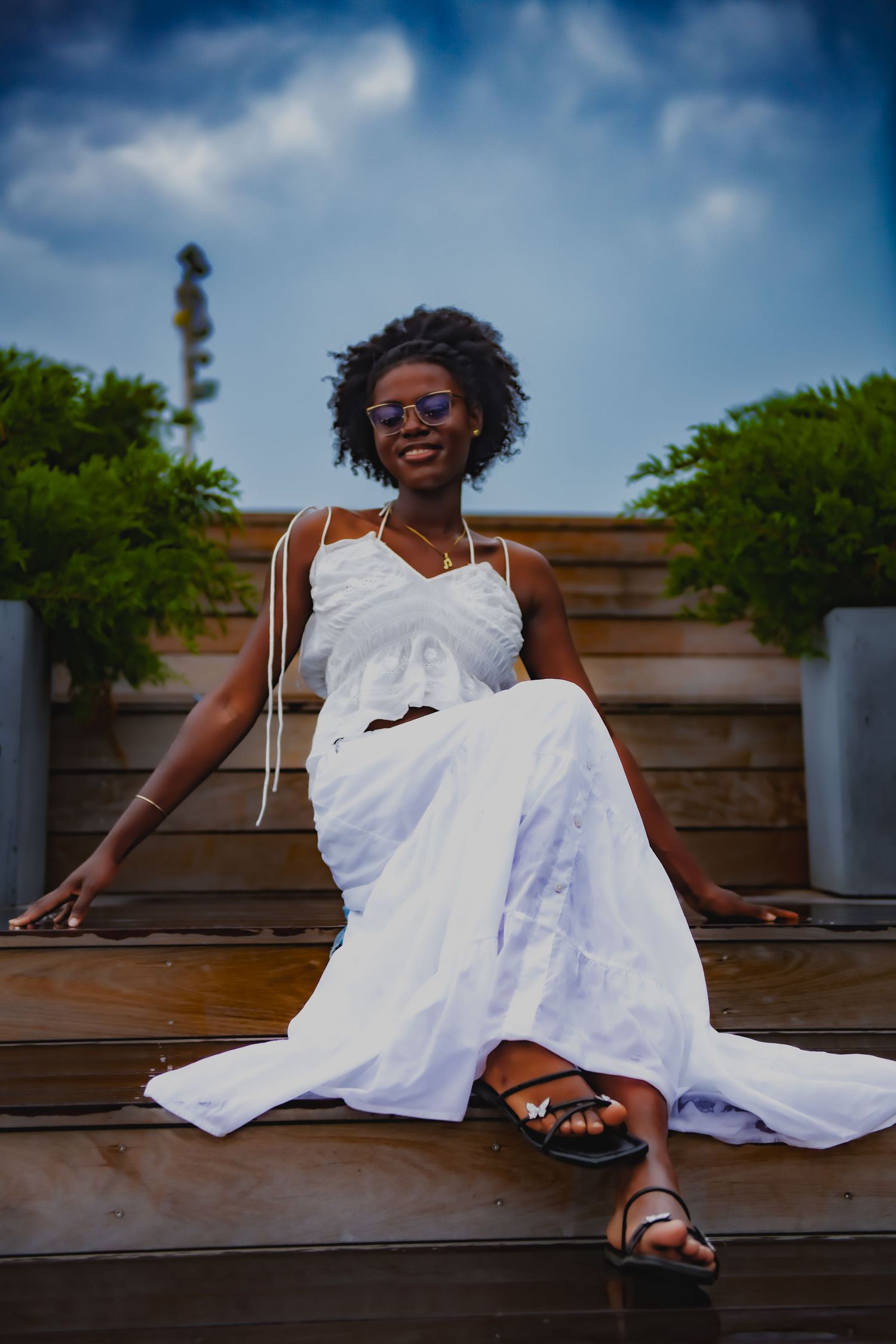 Relaxed pose in white dress on wooden steps under summer sky.