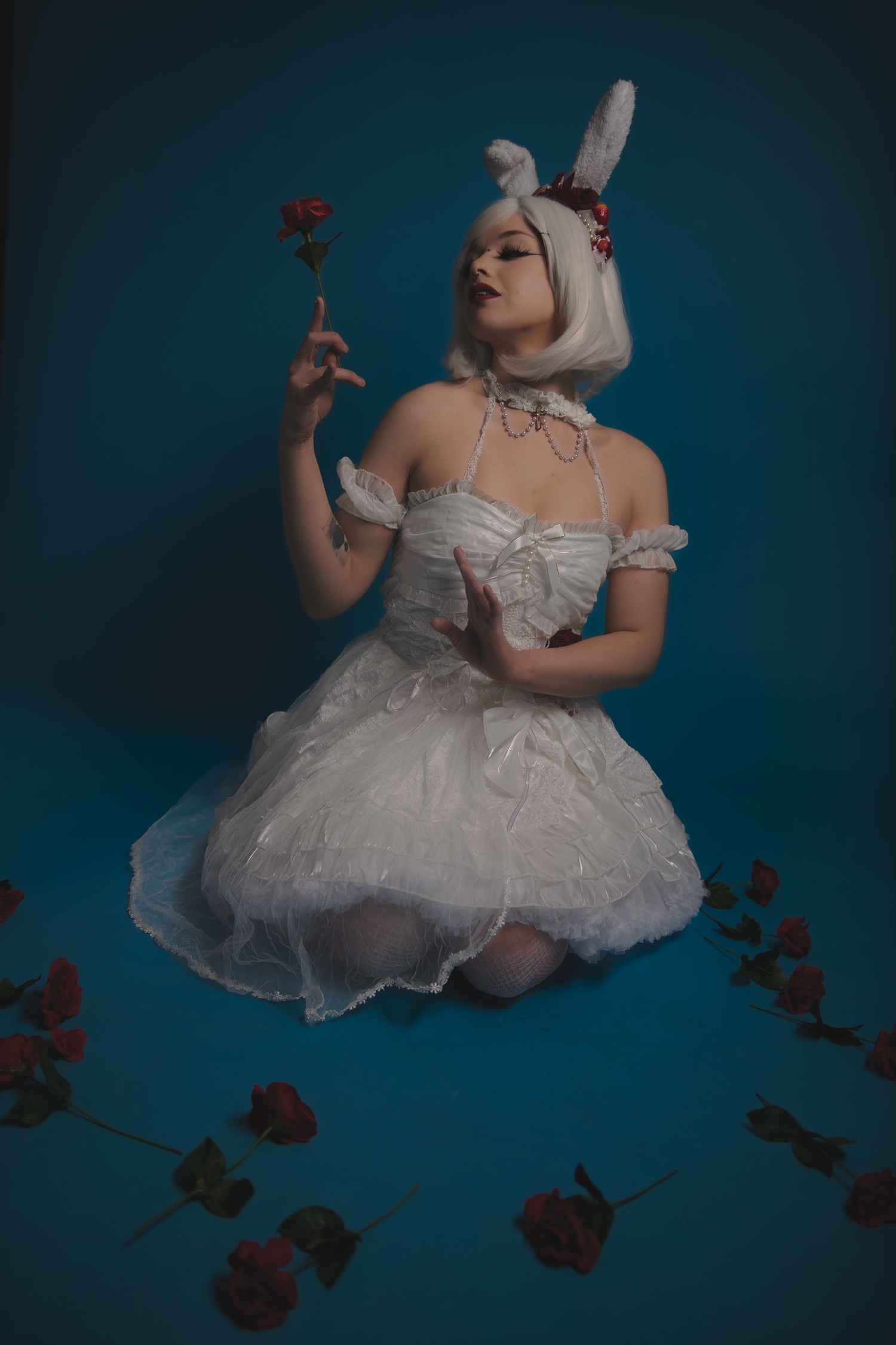 A person in a white ruffled dress poses artistically with red roses against a dark blue backdrop.