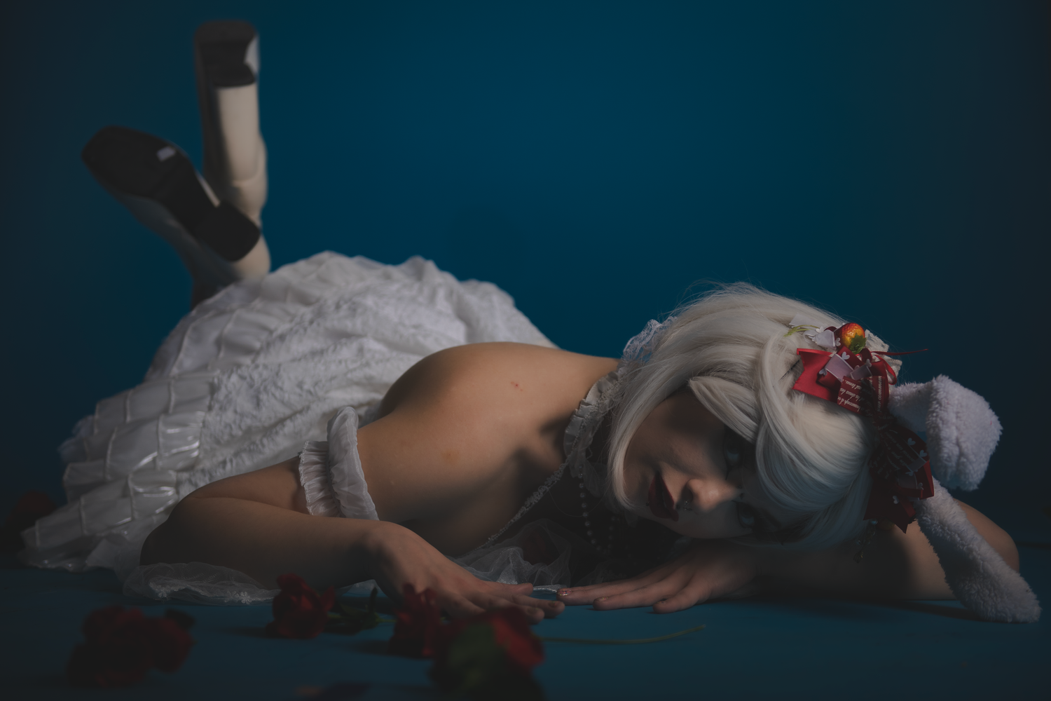 A person lies on white bedding in dim lighting with flowers scattered around them for a moody artistic photograph.