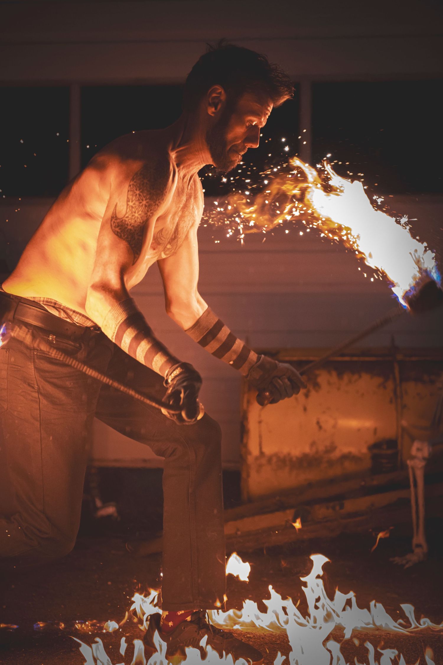 A fire performer breathes flames in a dramatic display while surrounded by sparks and fire on a dark stage.