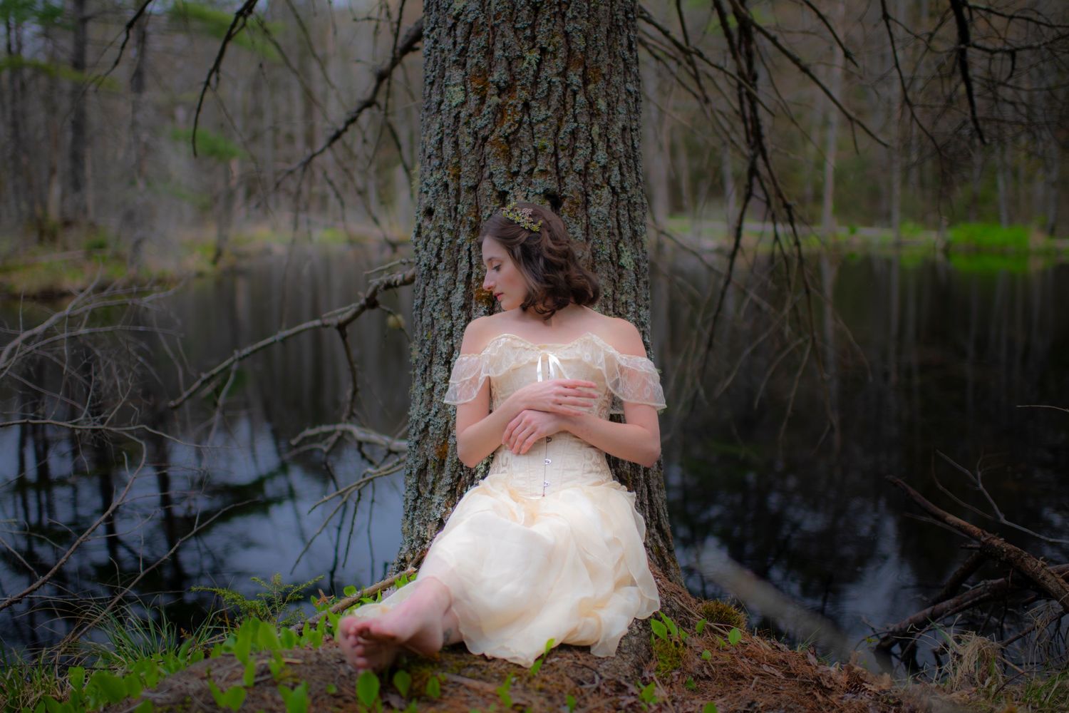 Person in ethereal dress sits against tree trunk by tranquil forest pond.