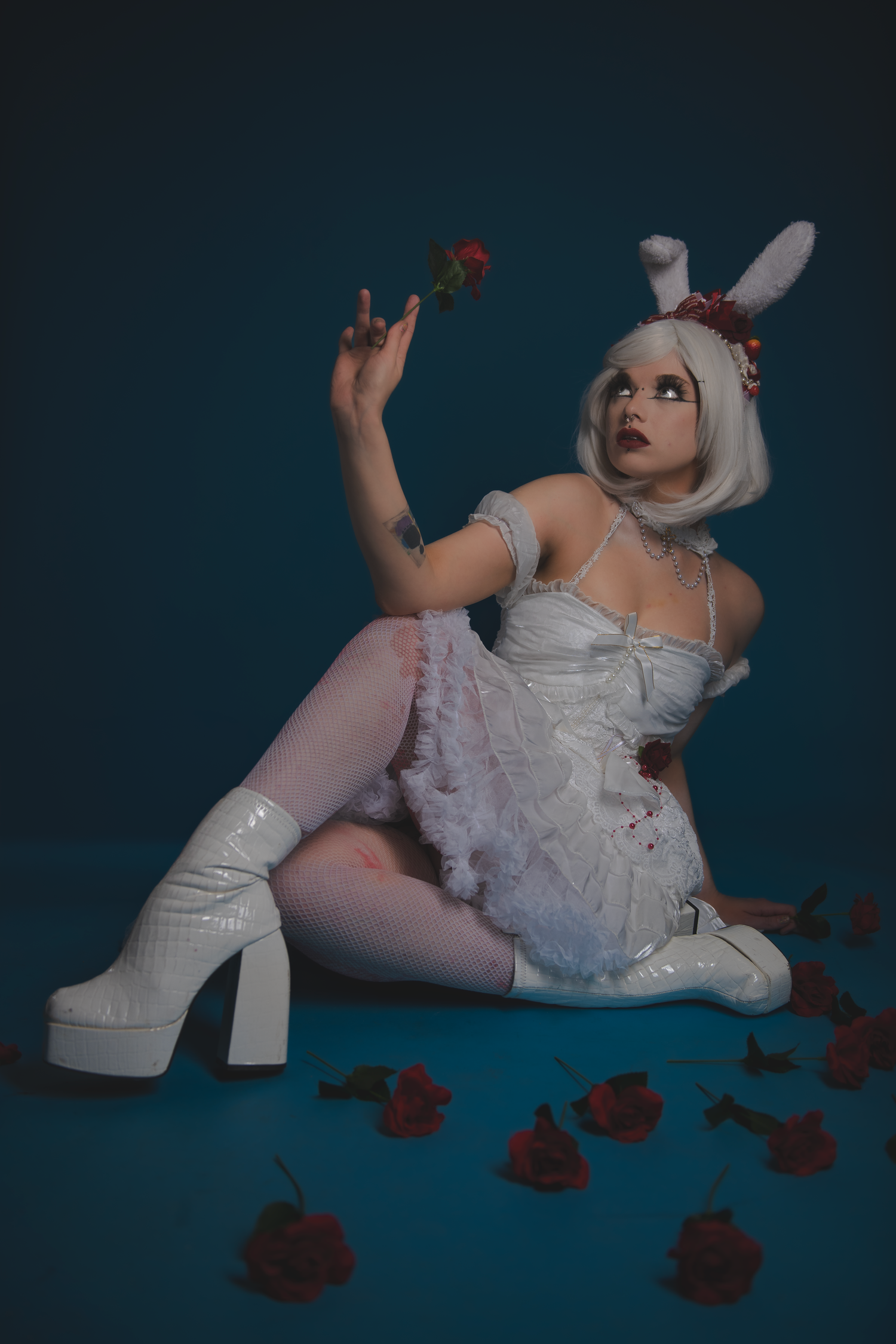 Stylized portrait with white platform boots, lace dress and scattered red roses on blue background.
