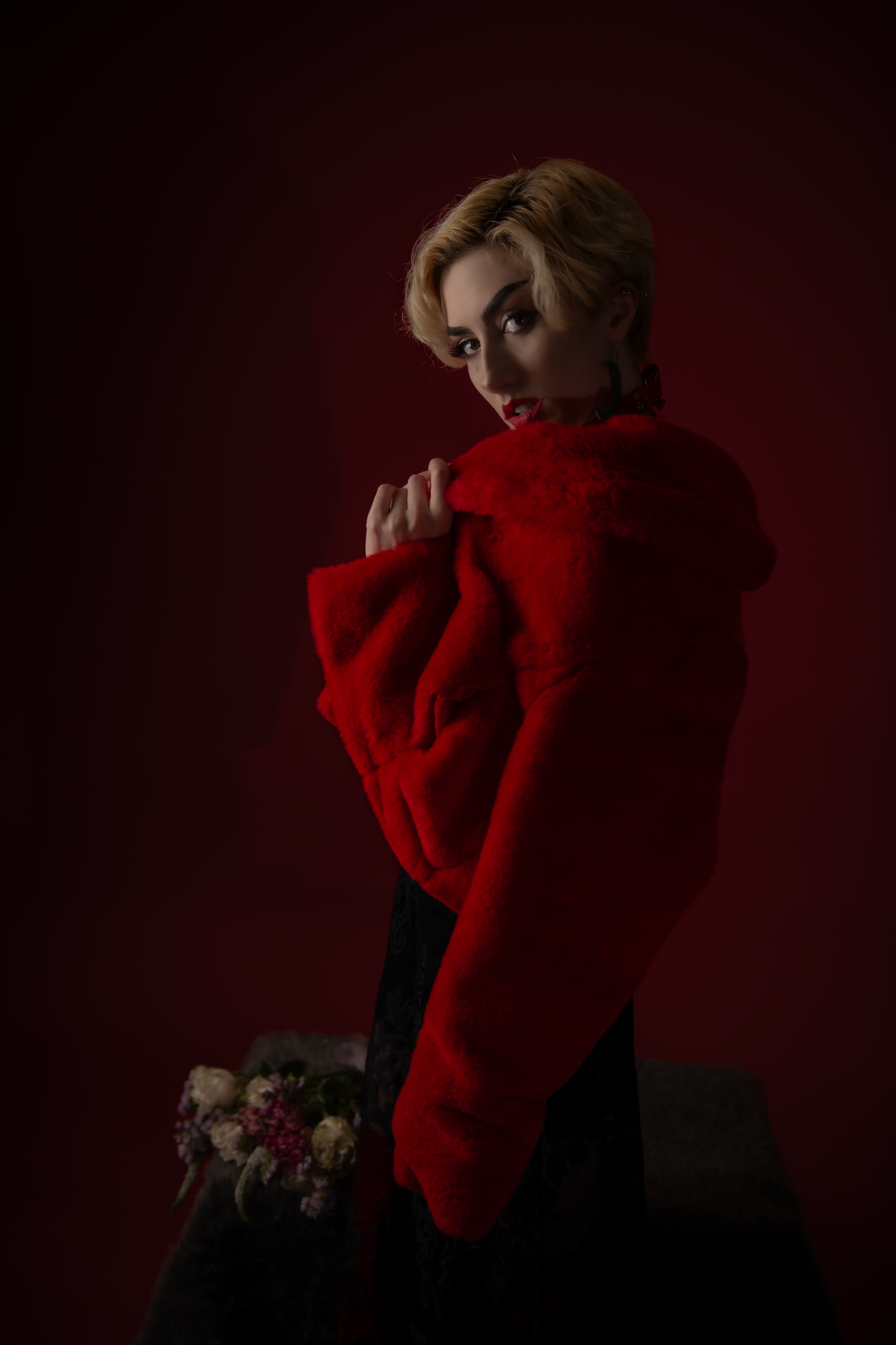 Dramatic portrait with figure in bright red sweater against dark burgundy background with subtle floral arrangement.