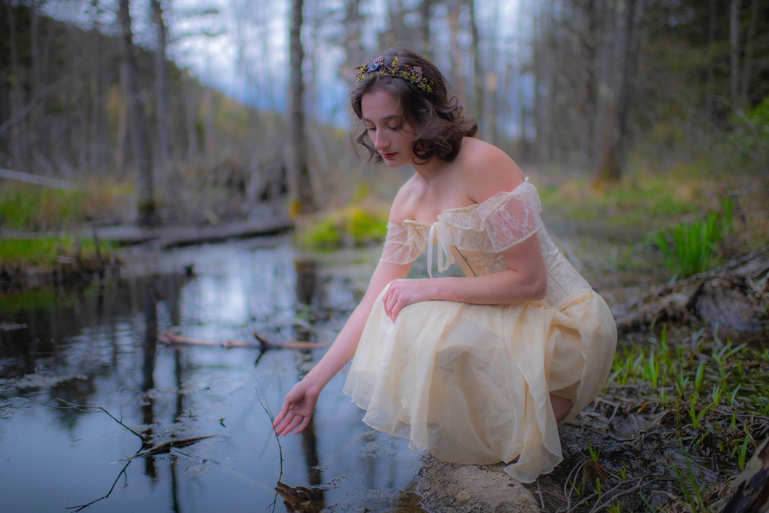 Figure in cream lace dress dips toes in forest pond during twilight.