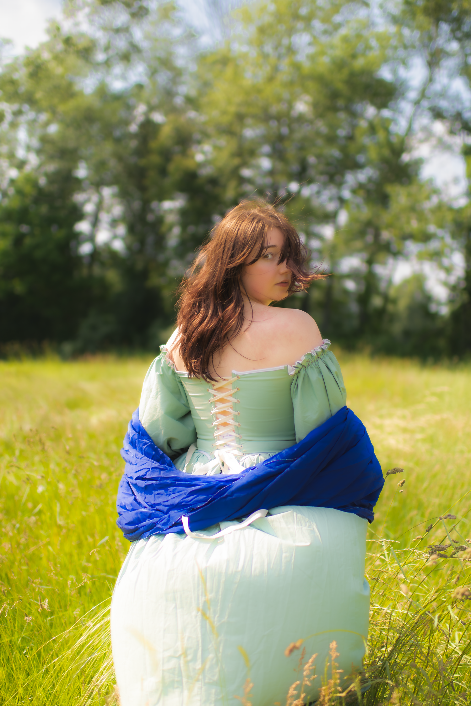 Person in mint green dress with flowing hair lying in sunny meadow with blue fabric against natural backdrop.