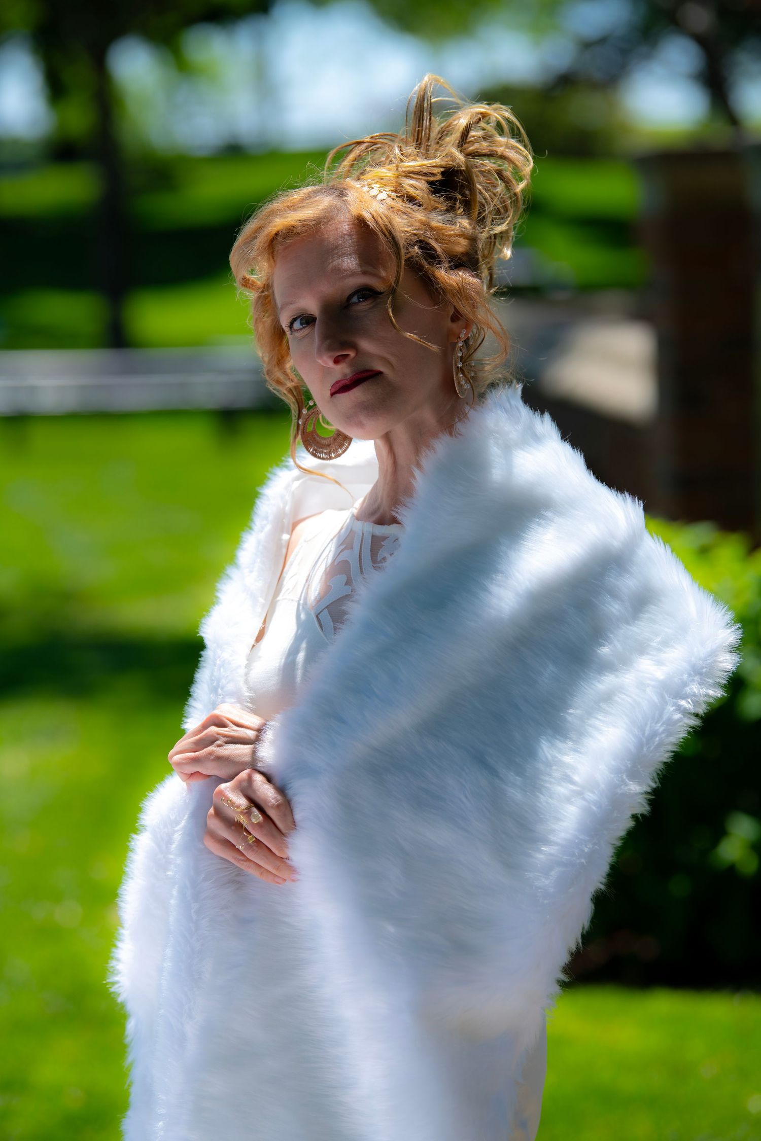 A person wearing a soft white fur wrap poses outdoors in natural lighting against a green background.