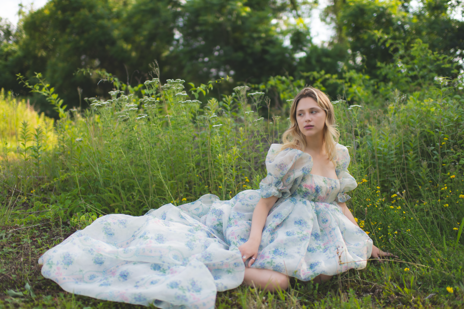 A person in a flowing floral dress sits gracefully in a lush green meadow surrounded by wildflowers and tall grass.